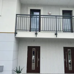 Elewacja domu z balkonem i dwoma wejściami, wykończona w pionowe pasy szarości i bieli, z ciemnymi oknami i drzwiami oraz czarną balustradą balkonu.