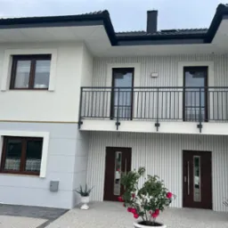 Dwupiętrowy dom z balkonem, elewacja w odcieniach szarości i bieli, z ciemnymi oknami i drzwiami, oraz kwiatami w donicach przed wejściem.