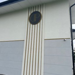 AT-BUD - Elewacja budynku z szarym pasem i pionowymi złotymi paskami, okrągłe logo firmy i kamera monitoringu