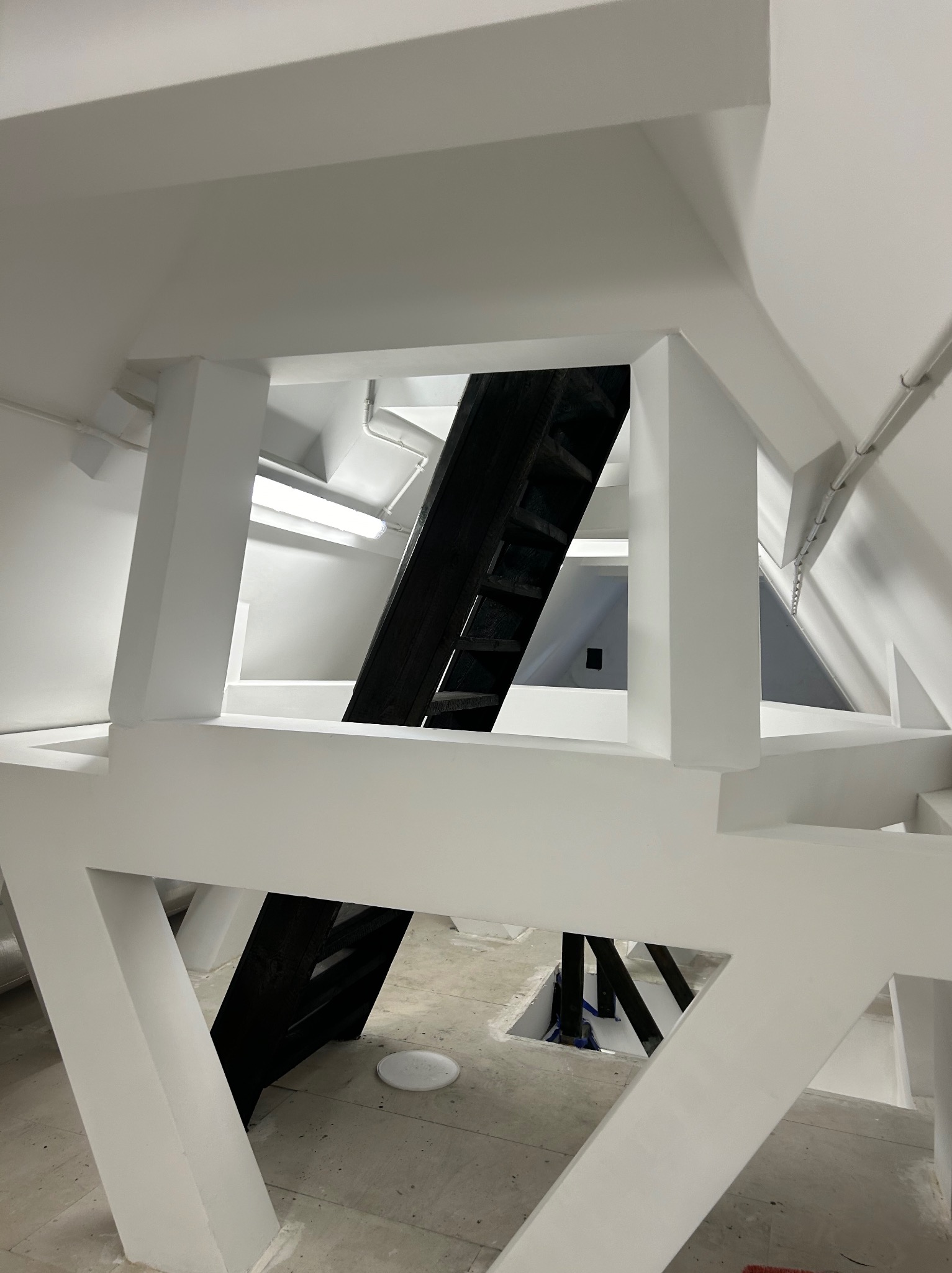 Wnętrze poddasza z białą konstrukcją nośną i czarnymi schodami, widok z dołu, surowy beton na podłodze, oświetlenie ledowe.