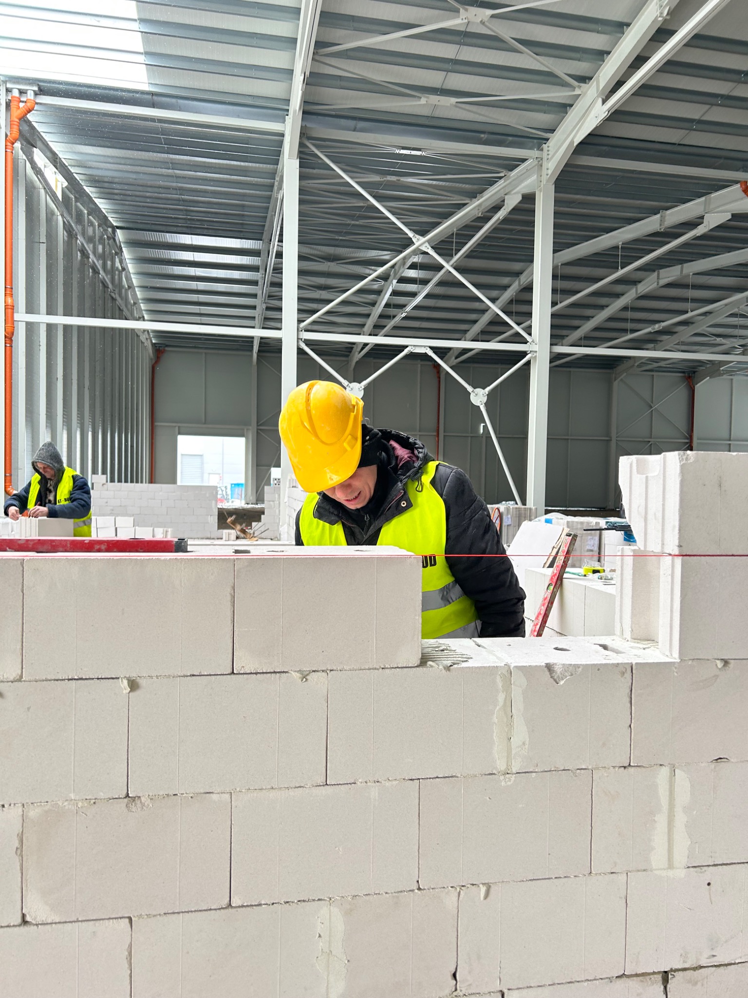 Murarz w żółtym kasku i kamizelce, w trakcie murowania ściany z białych bloczków betonu komórkowego wewnątrz hali, z widocznym sznurkiem murarskim i poziomnicą, drugi pracownik w tle.
