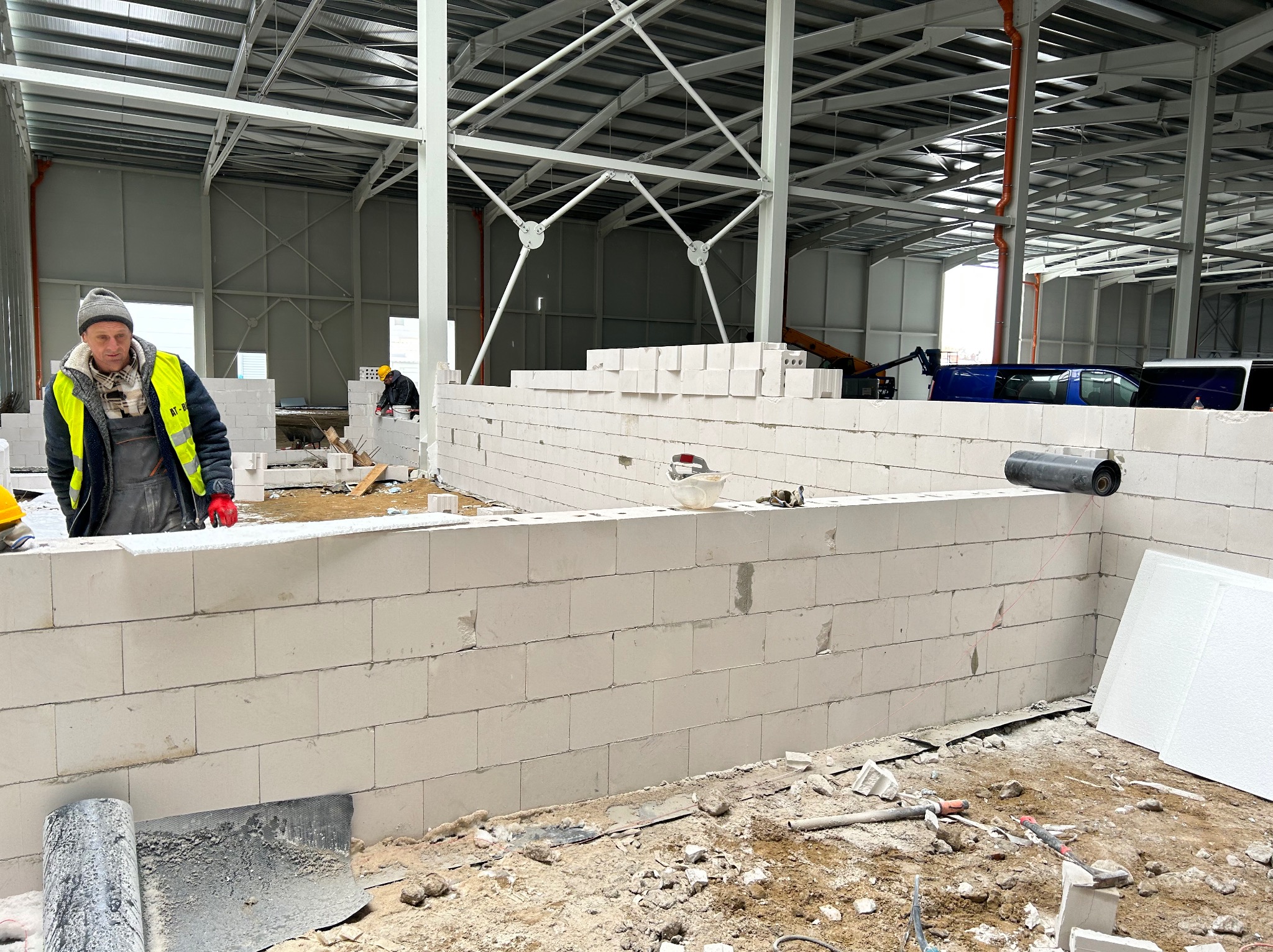 Wznoszenie ściany z betonu komórkowego w hali przemysłowej, widoczny pracownik w odblaskowej kamizelce i czapce, w tle stalowa konstrukcja dachu i zaparkowane busy