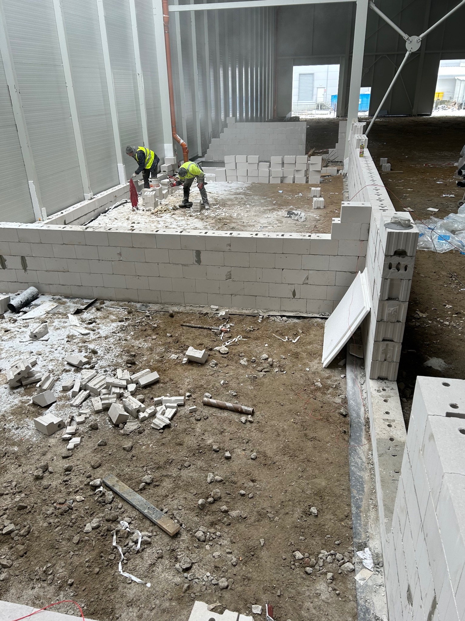 Wznoszenie ścian z betonu komórkowego Ytong wewnątrz hali, dwóch robotników w kamizelkach odblaskowych przycina bloczki, widoczne porozrzucane fragmenty materiału i narzędzia.