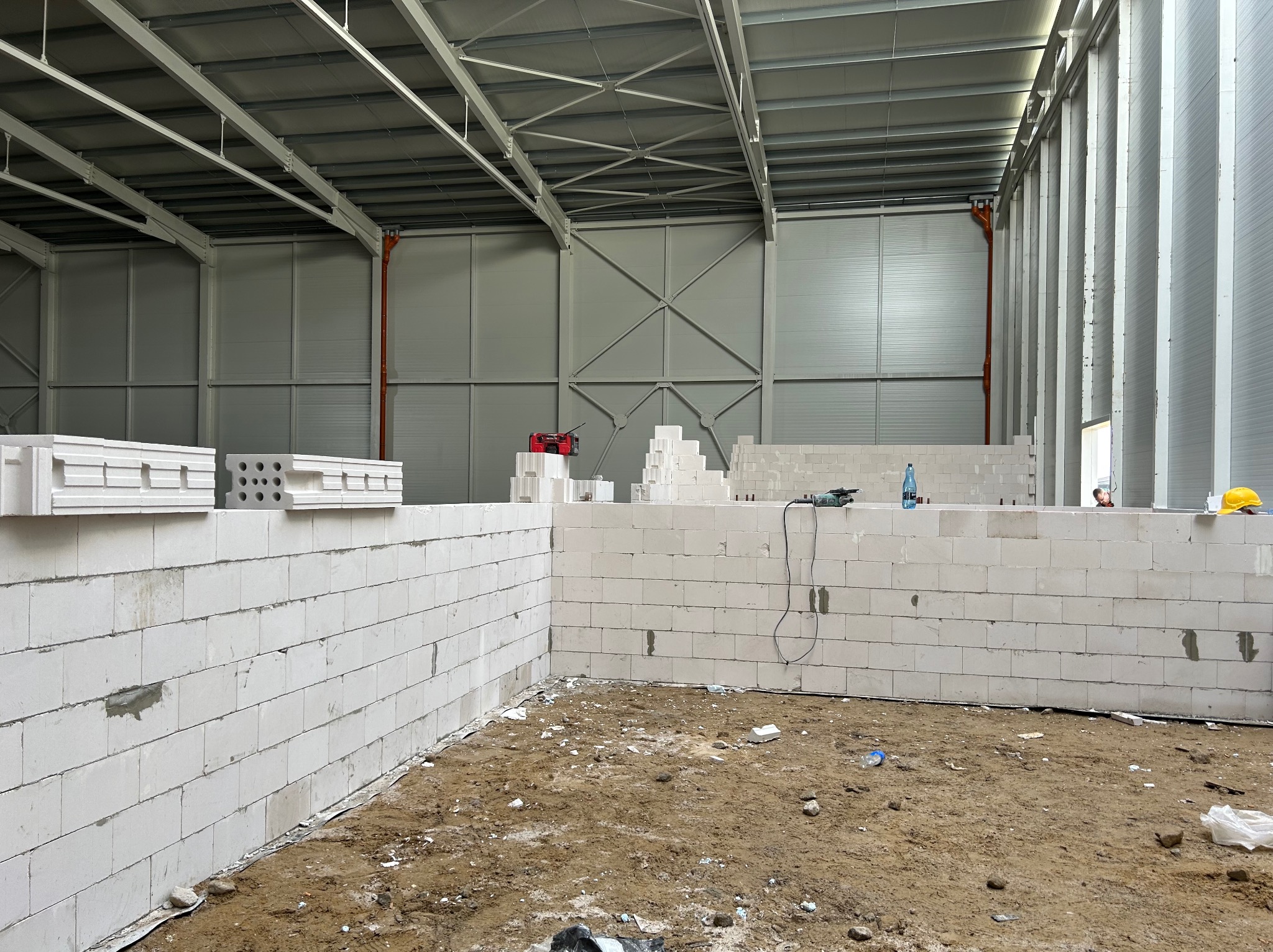 Wnętrze hali w trakcie budowy ścian z białych bloczków, widoczne narzędzia, resztki materiałów budowlanych na ziemi, konstrukcja dachu hali.