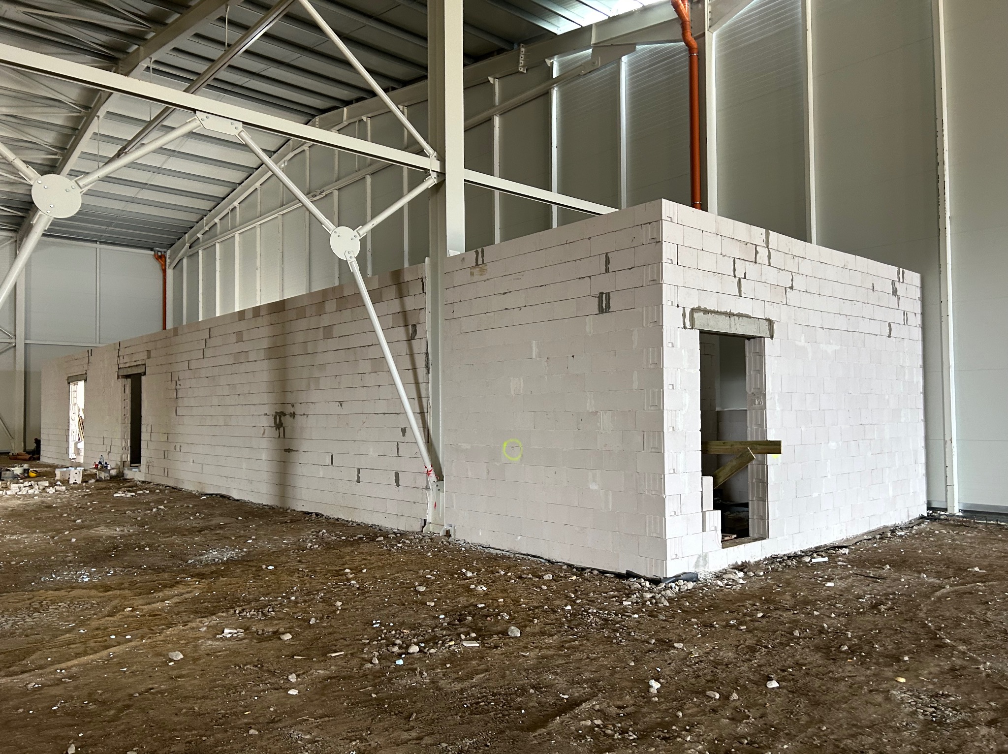 Wnętrze hali w trakcie budowy ścian działowych z białych bloczków, widoczne otwory drzwiowe z drewnianymi stemplami, surowa podłoga i metalowa konstrukcja dachu.