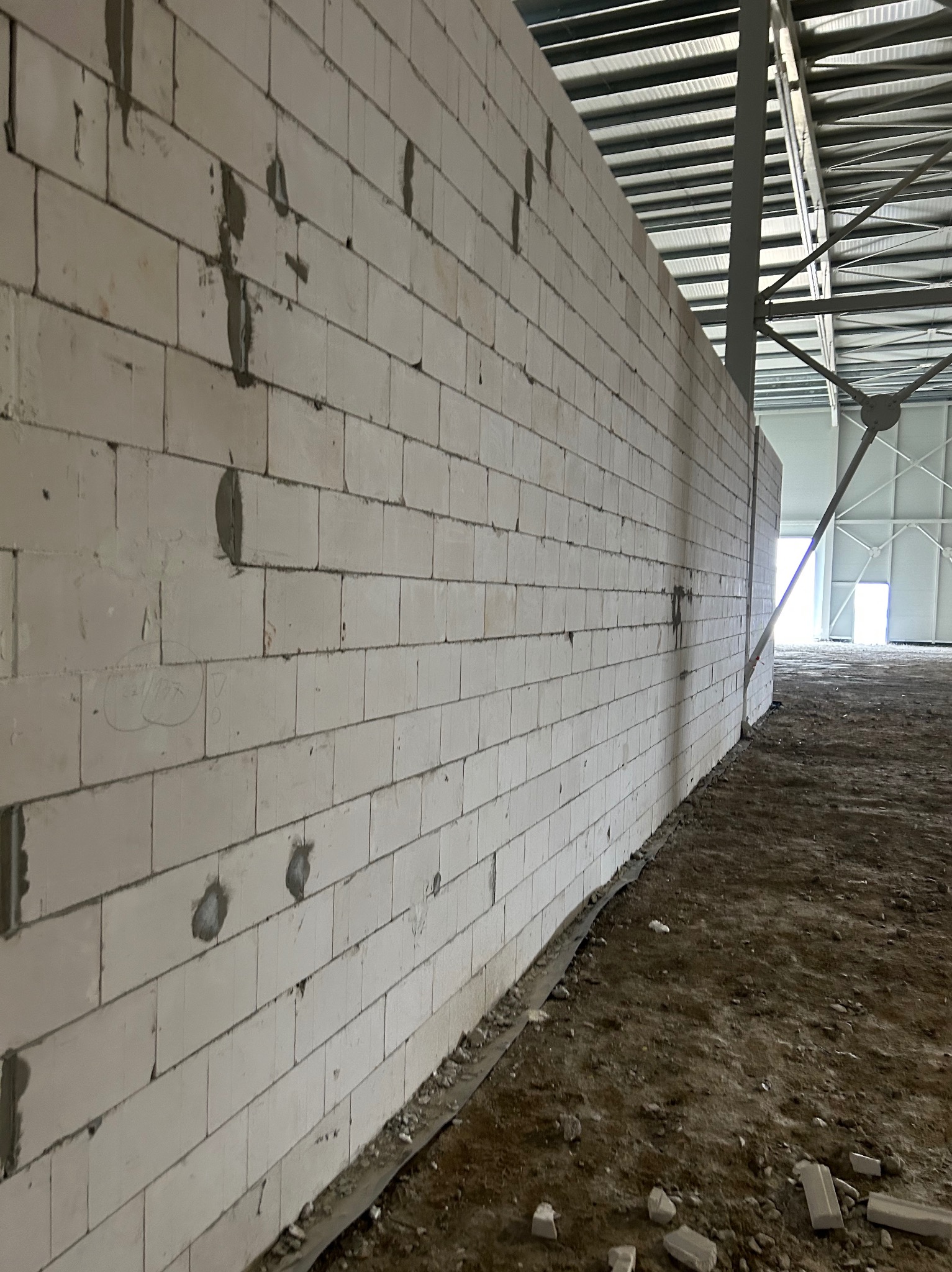 Ściana z białych bloczków betonowych w surowym stanie, z widocznymi śladami zaprawy i niedoskonałościami, w hali z metalową konstrukcją dachu i ziemistą podłogą.