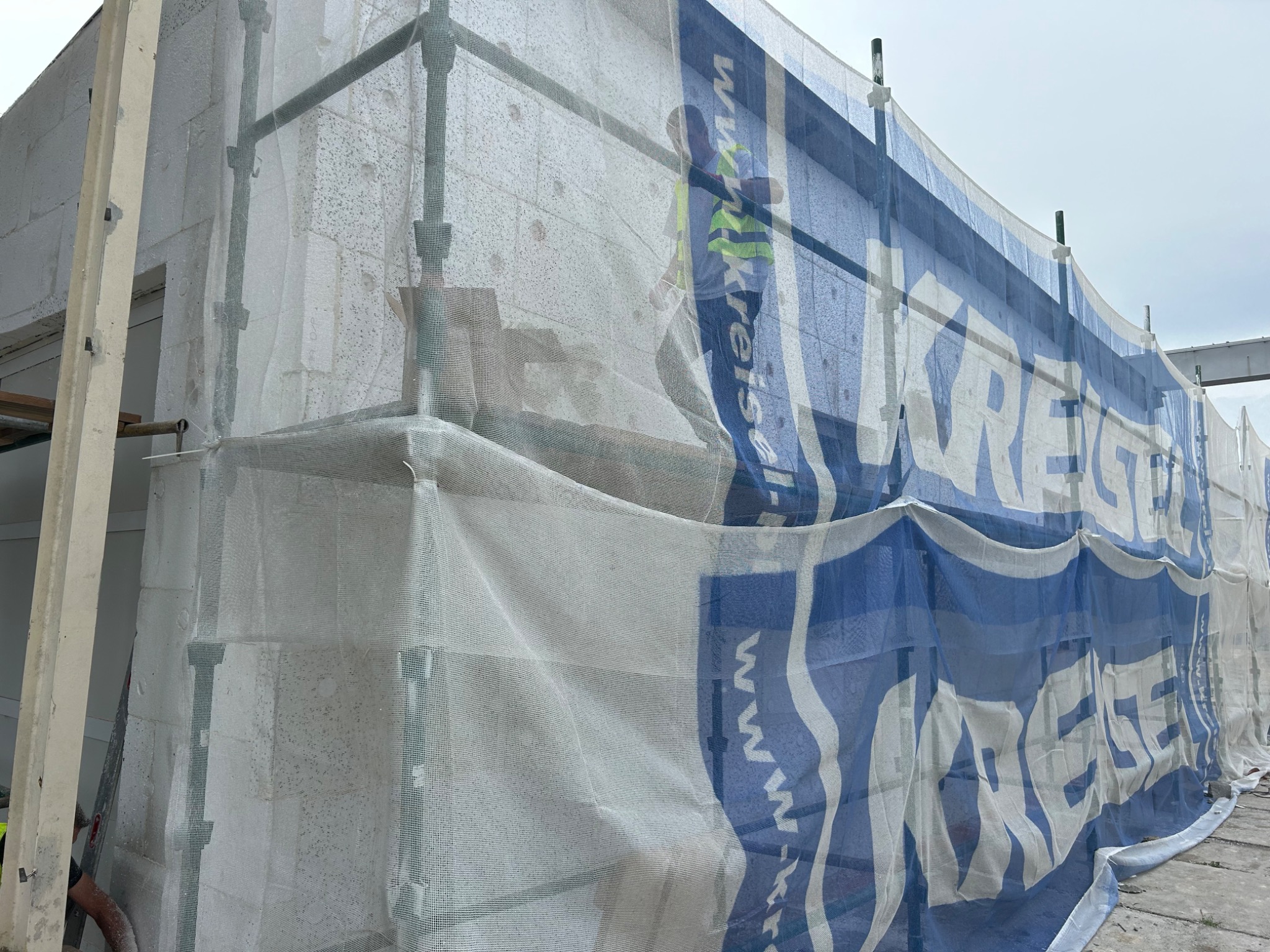 Budynek w trakcie ocieplania elewacji, widoczne rusztowania osłonięte siatką z reklamą, częściowo zasłaniającą ściany z bloczków betonu komórkowego.