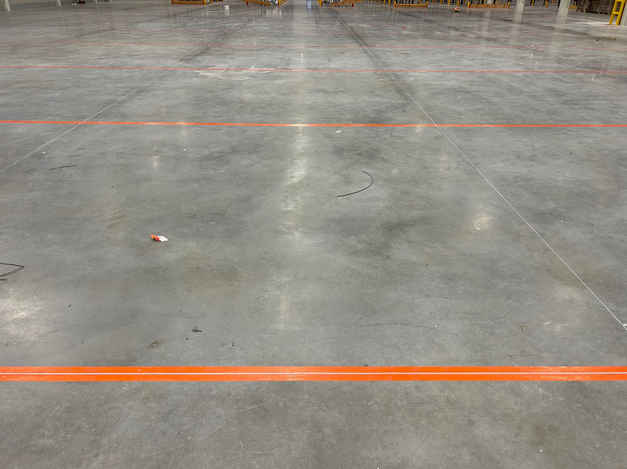 Szara, betonowa posadzka hali magazynowej z wyznaczonymi pomarańczowymi liniami poziomymi, widoczne drobne zabrudzenia i resztki materiałów budowlanych.
