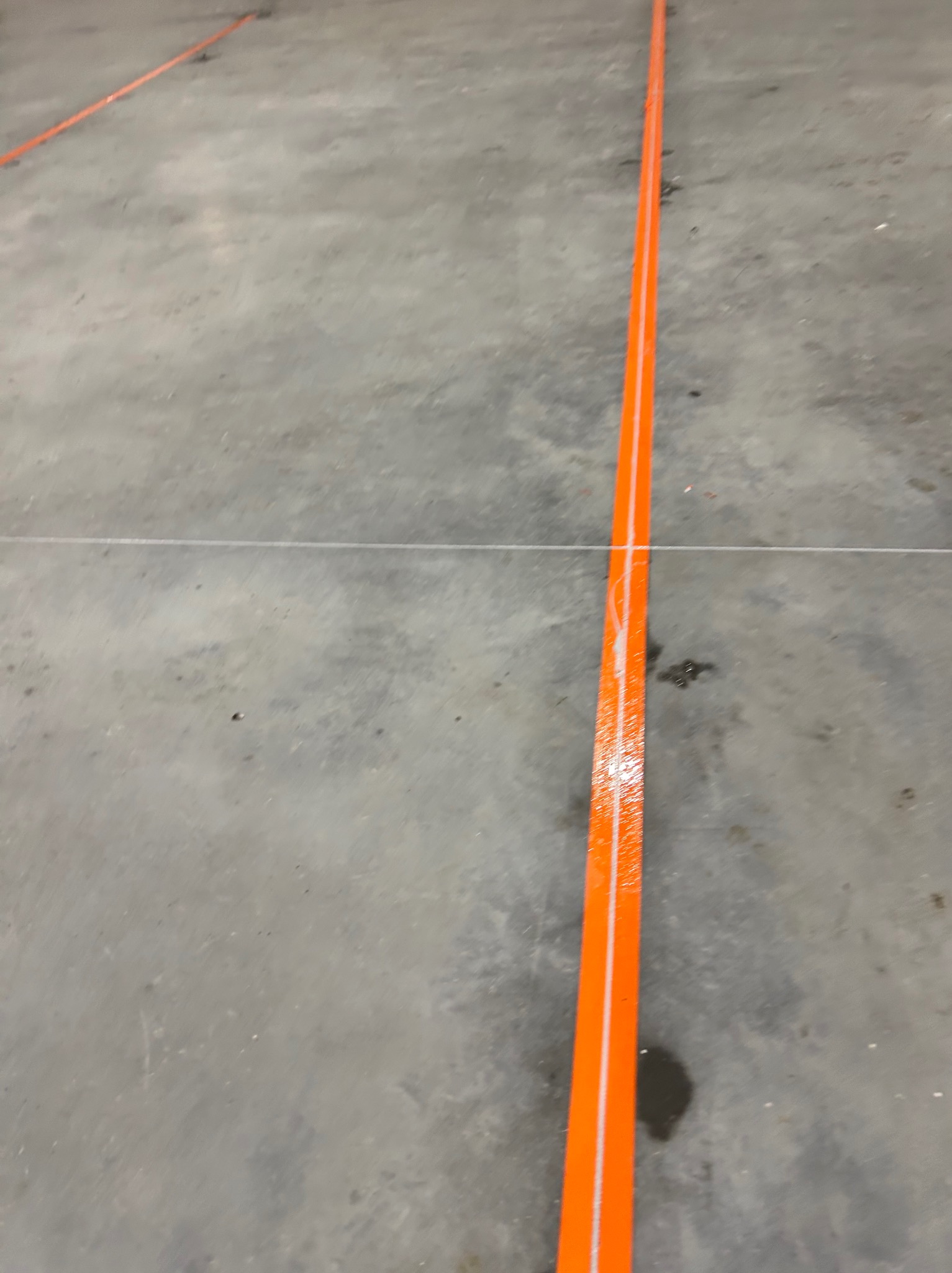 Świeżo pomalowana, jaskrawo pomarańczowa linia na szarej, betonowej podłodze, z widocznymi śladami niedokładności malowania i dodatkową, cienką linią wyznaczającą jej położenie.