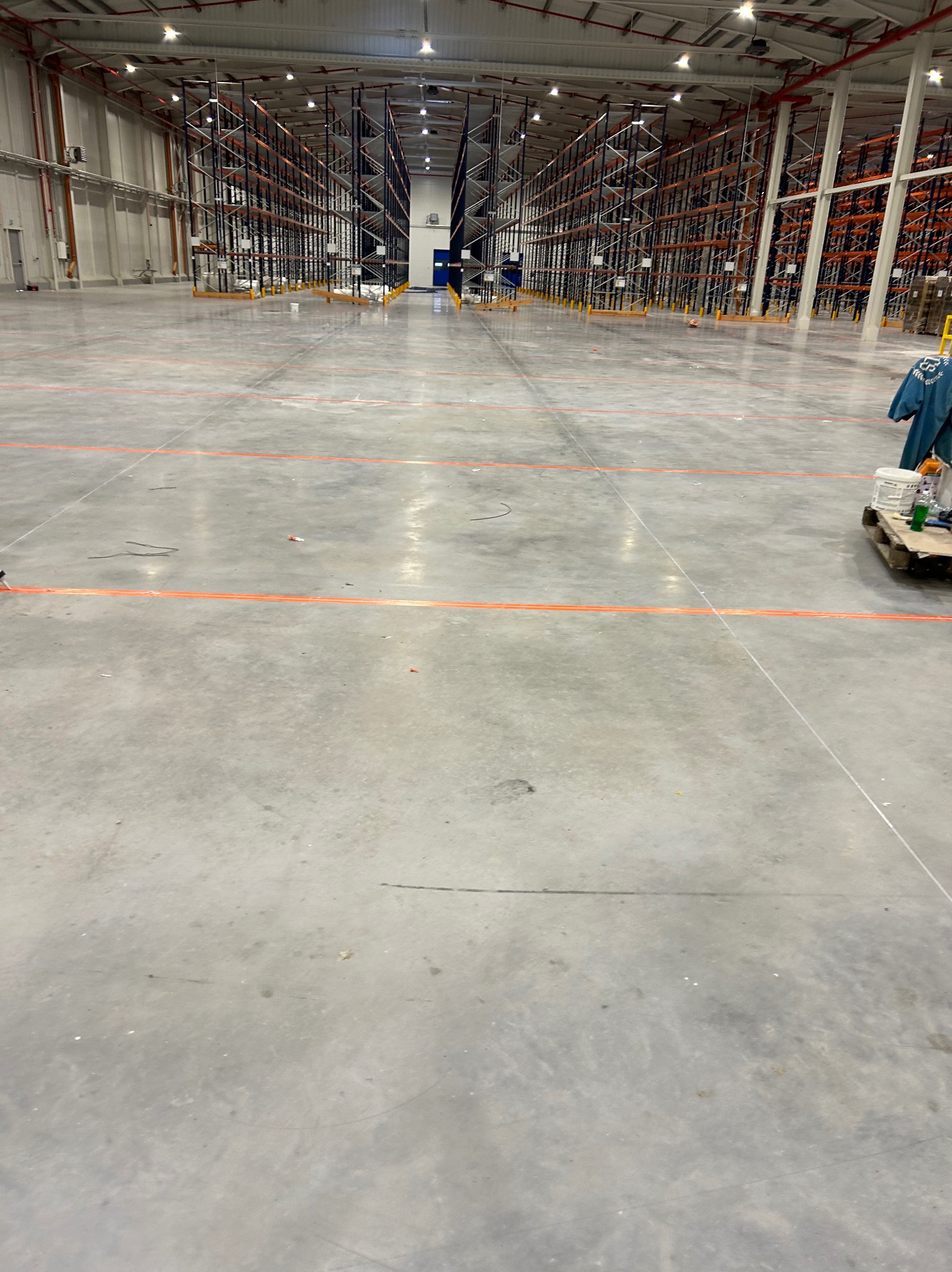 Wnętrze pustego magazynu z betonową podłogą z pomarańczowymi liniami, metalowymi regałami w tle i paletą z farbami oraz ubraniem roboczym po prawej stronie.