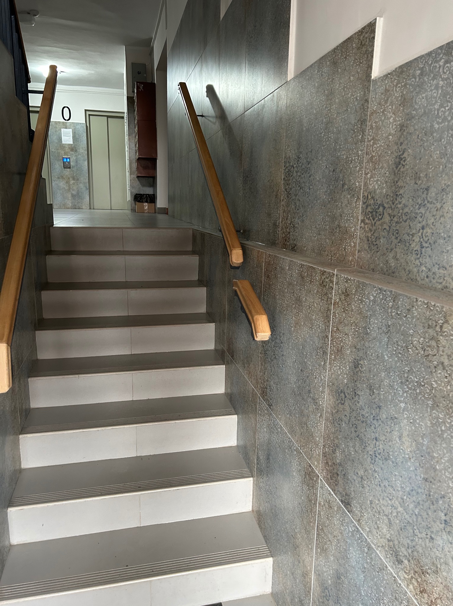 Klatka schodowa w budynku z wykończonymi schodami z płytek i drewnianą balustradą, widok na korytarz z windą.