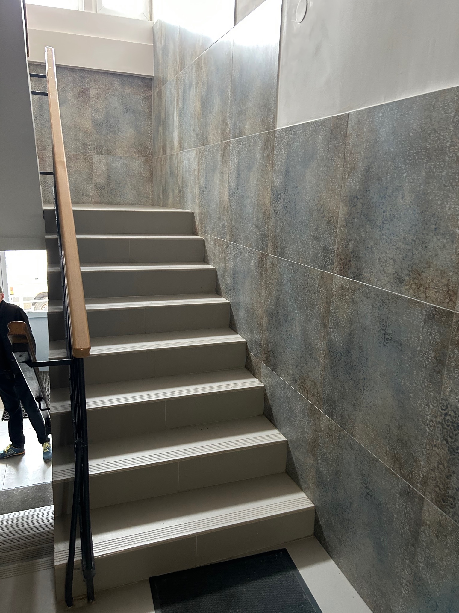 Wykończone schody wewnętrzne z szarobrązowymi płytkami dekoracyjnymi na ścianie i jasnymi stopniami, widoczny fragment poręczy i osoba w tle.