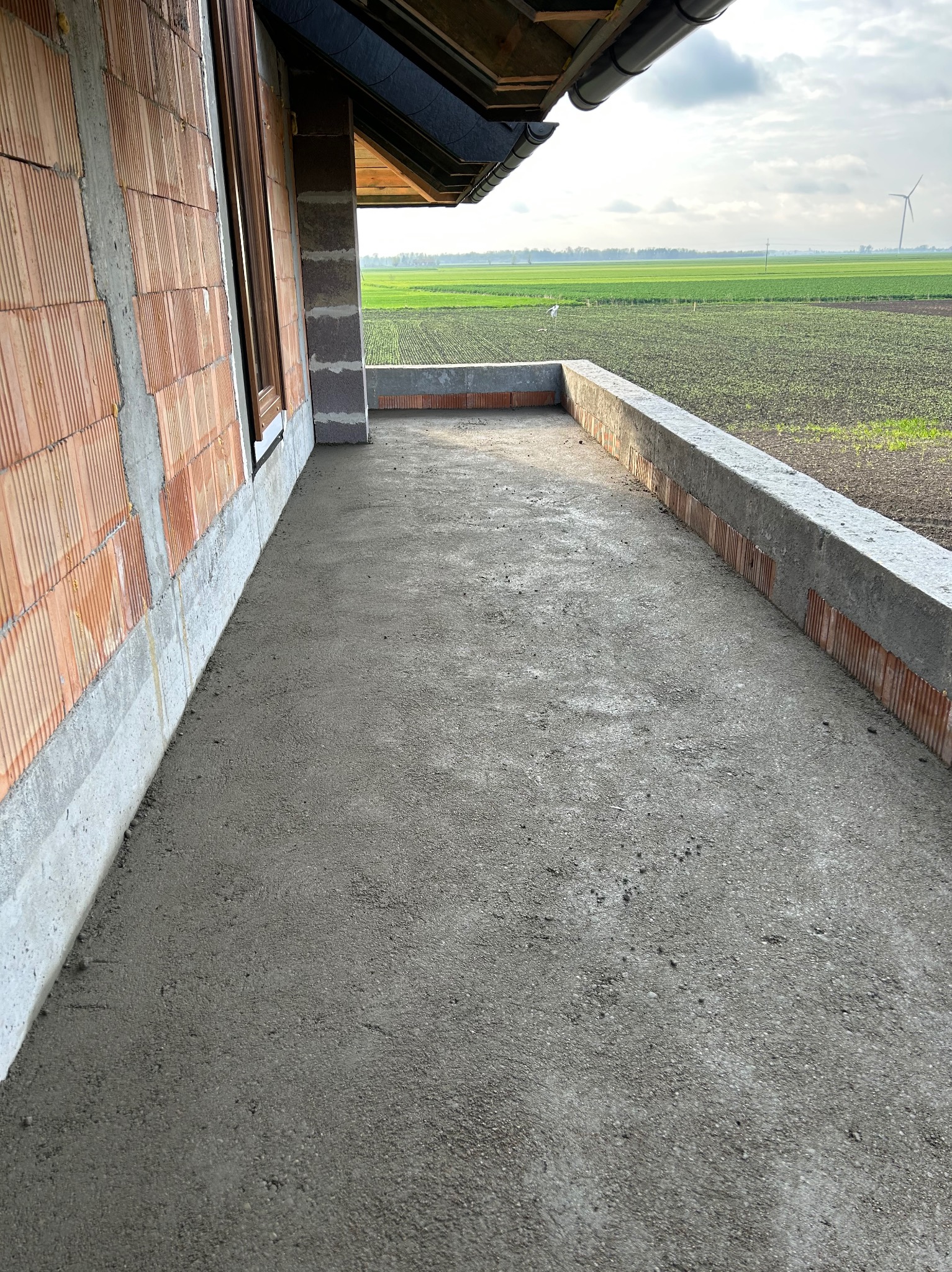 Świeżo wylany beton na balkonie domu w stanie surowym otwartym, widok na pola i turbinę wiatrową w tle.