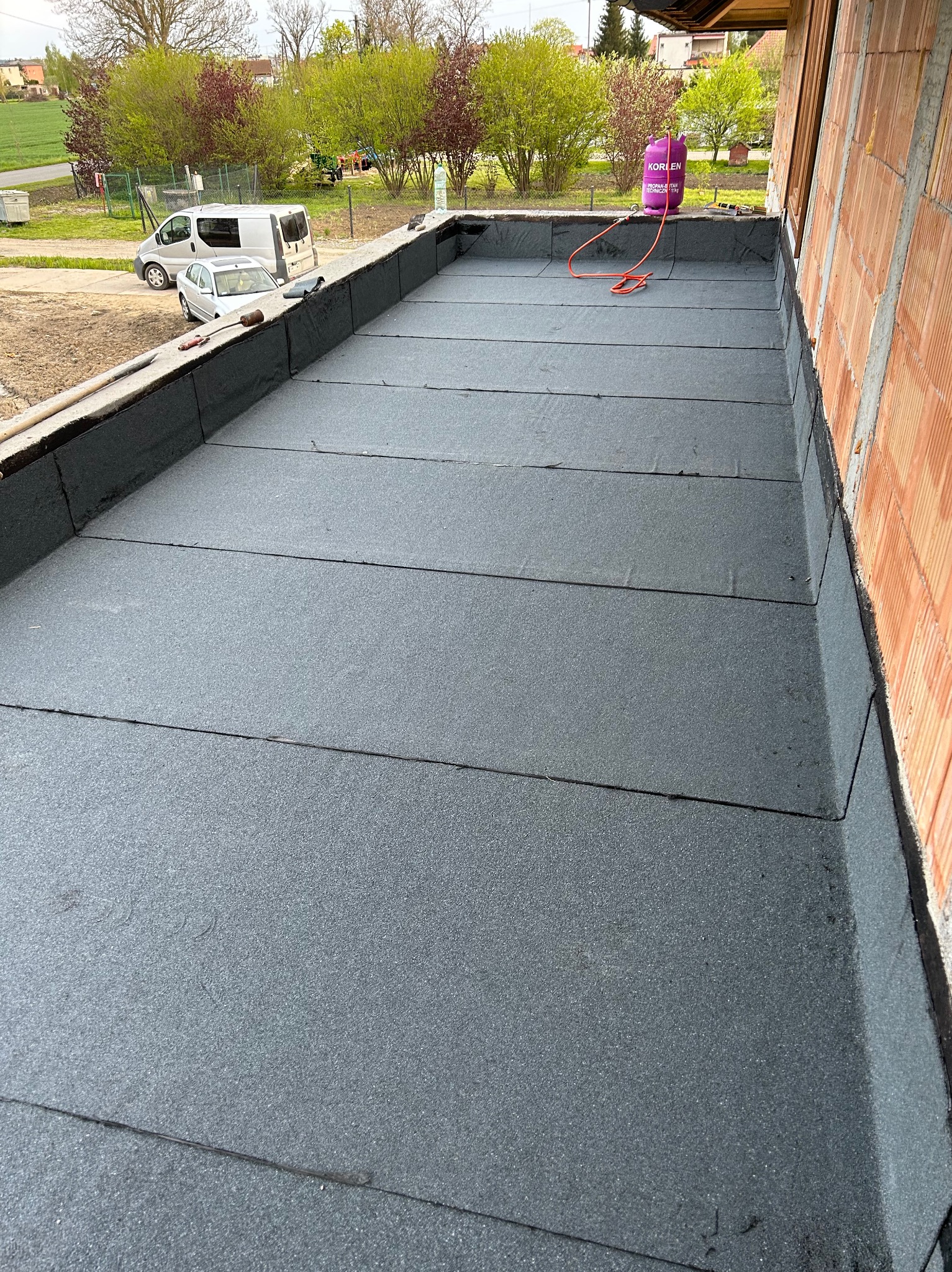 Wykończony płaski dach pokryty papą termozgrzewalną, widoczny gazowy palnik z butlą propan-butan, ściana budynku w tle.