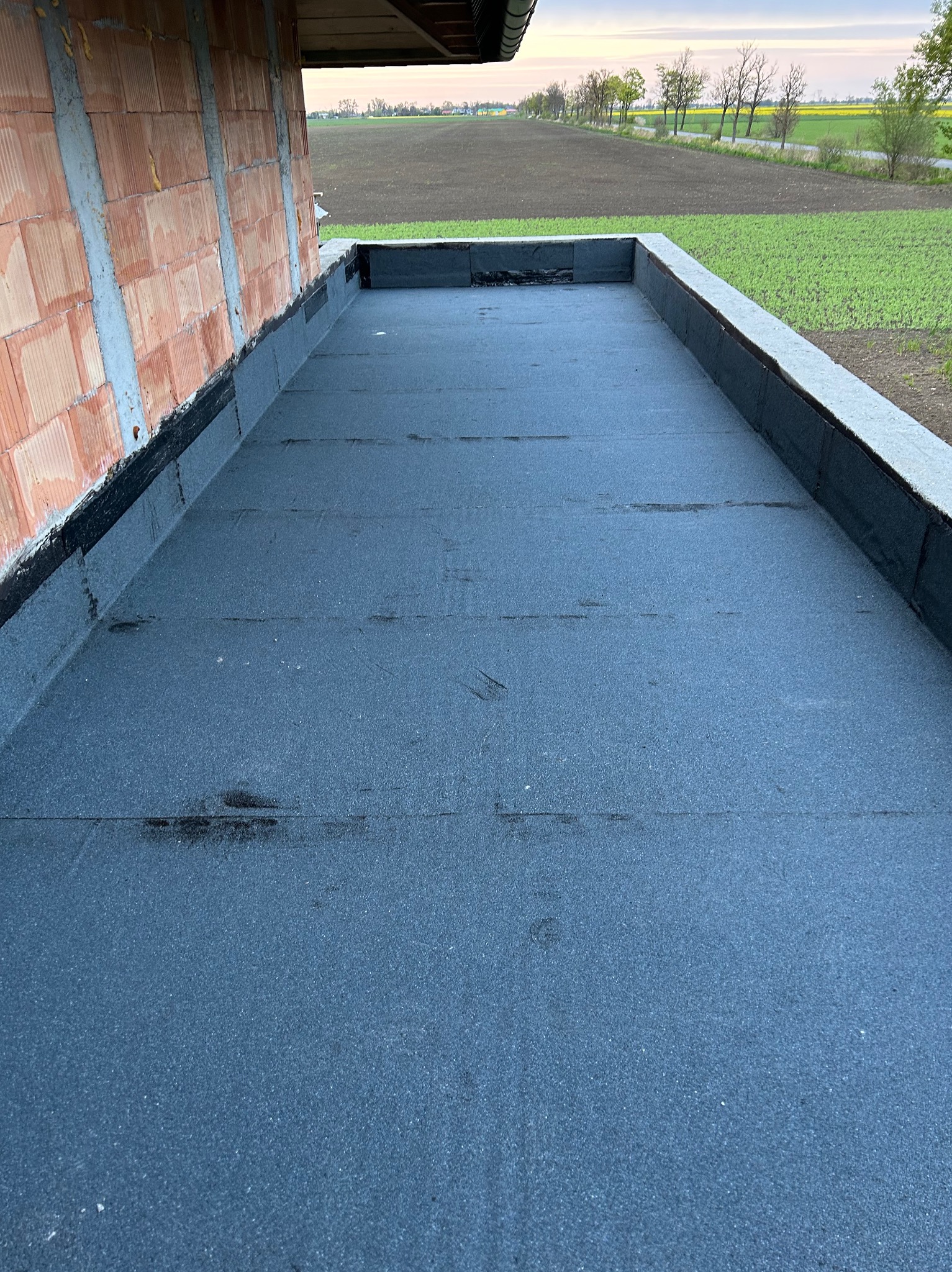 Wykończony płaski dach z papy termozgrzewalnej, widok z góry, surowa ściana budynku w tle, za nią pola i drzewa.