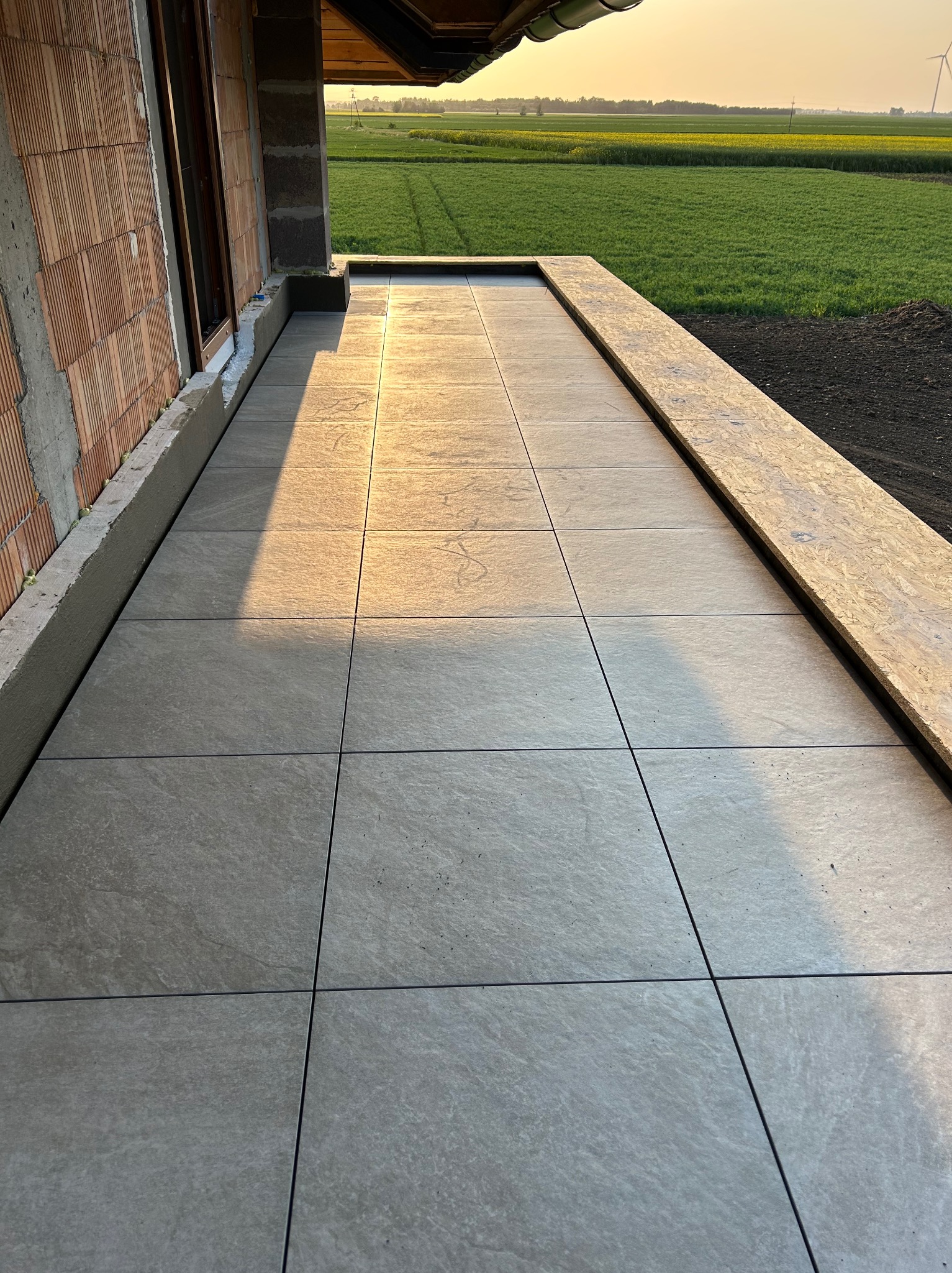 Świeżo ułożone szare płytki na tarasie domu w trakcie budowy, widoczne cegły i ocieplenie, w tle pole i zachodzące słońce.