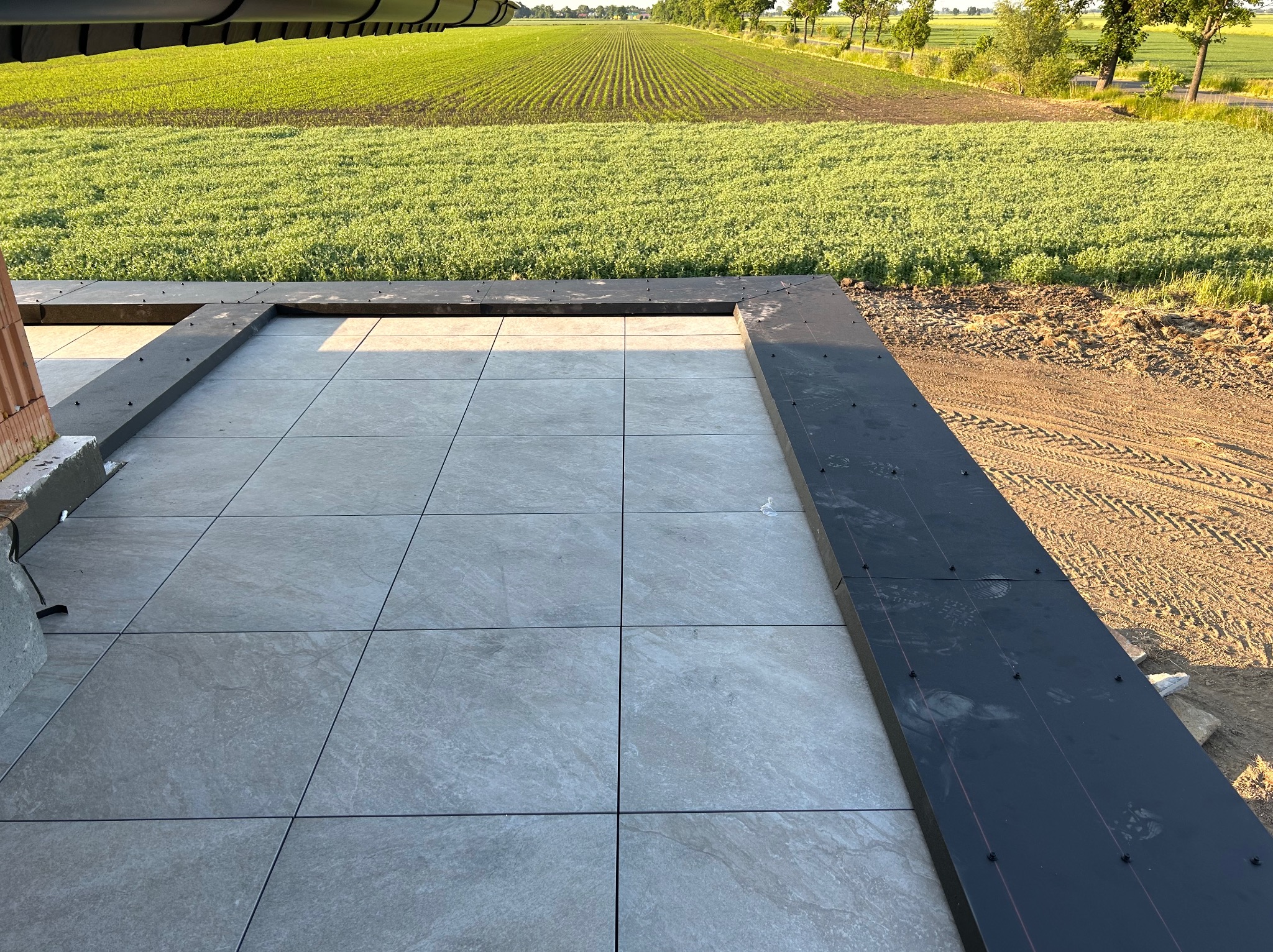 Szare płytki tarasowe ułożone na zewnątrz budynku, z widokiem na pole uprawne w tle, czarne obramowanie z widocznymi śladami butów.
