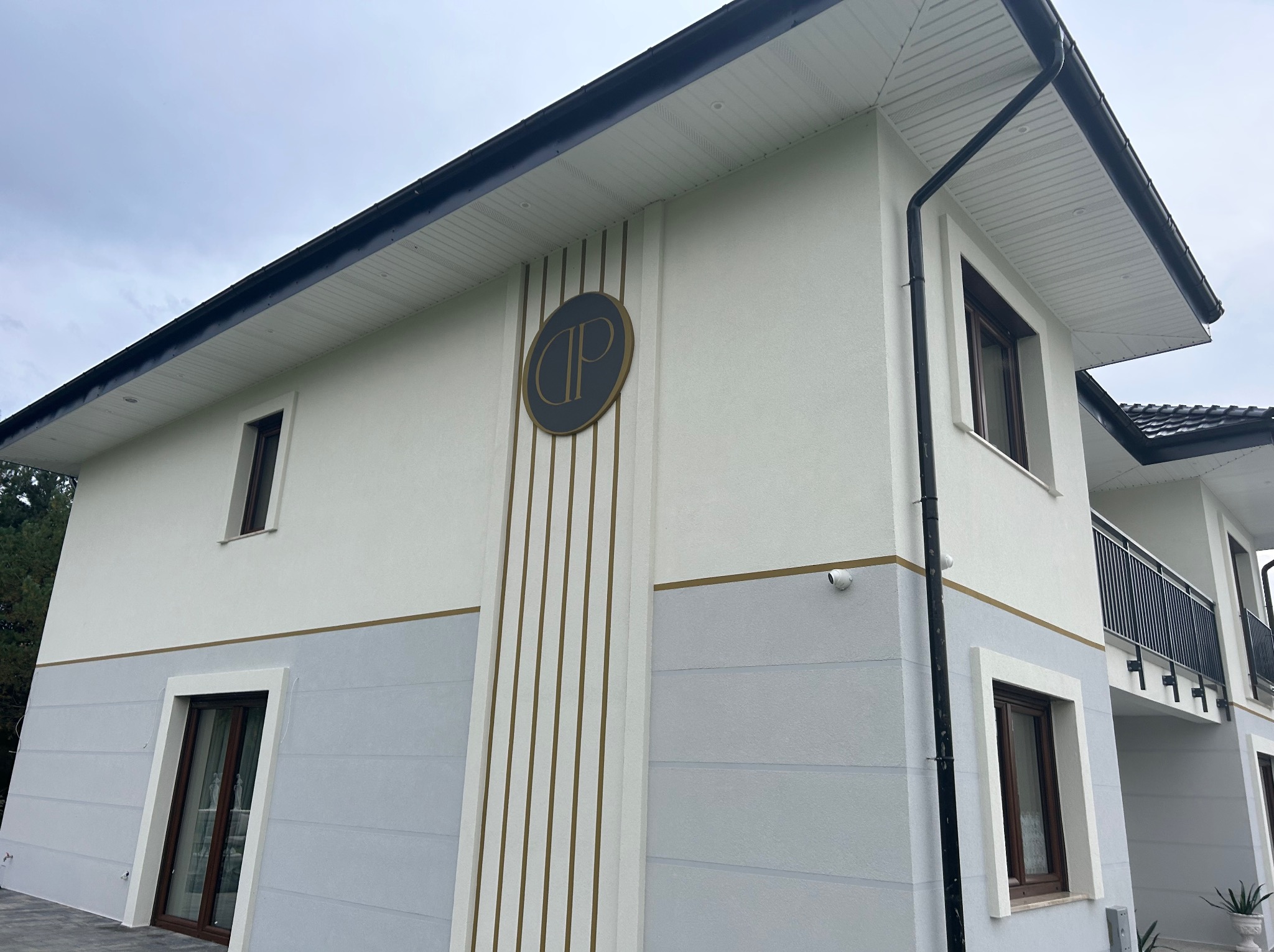 Dwukondygnacyjny budynek z jasną elewacją, podzieloną poziomym pasem na szarą i białą część, z widocznym logo firmy w kształcie okręgu z literami na elewacji i czarnym dachem.