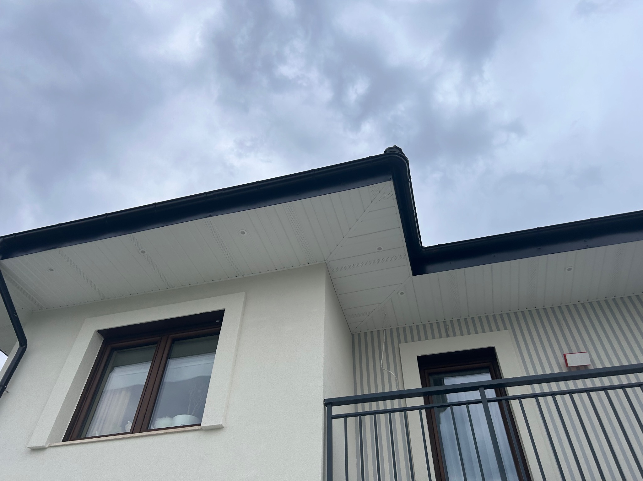 Fragment elewacji domu jednorodzinnego z widocznym orynnowaniem, podbitką dachową i balkonem z metalową balustradą na tle pochmurnego nieba.