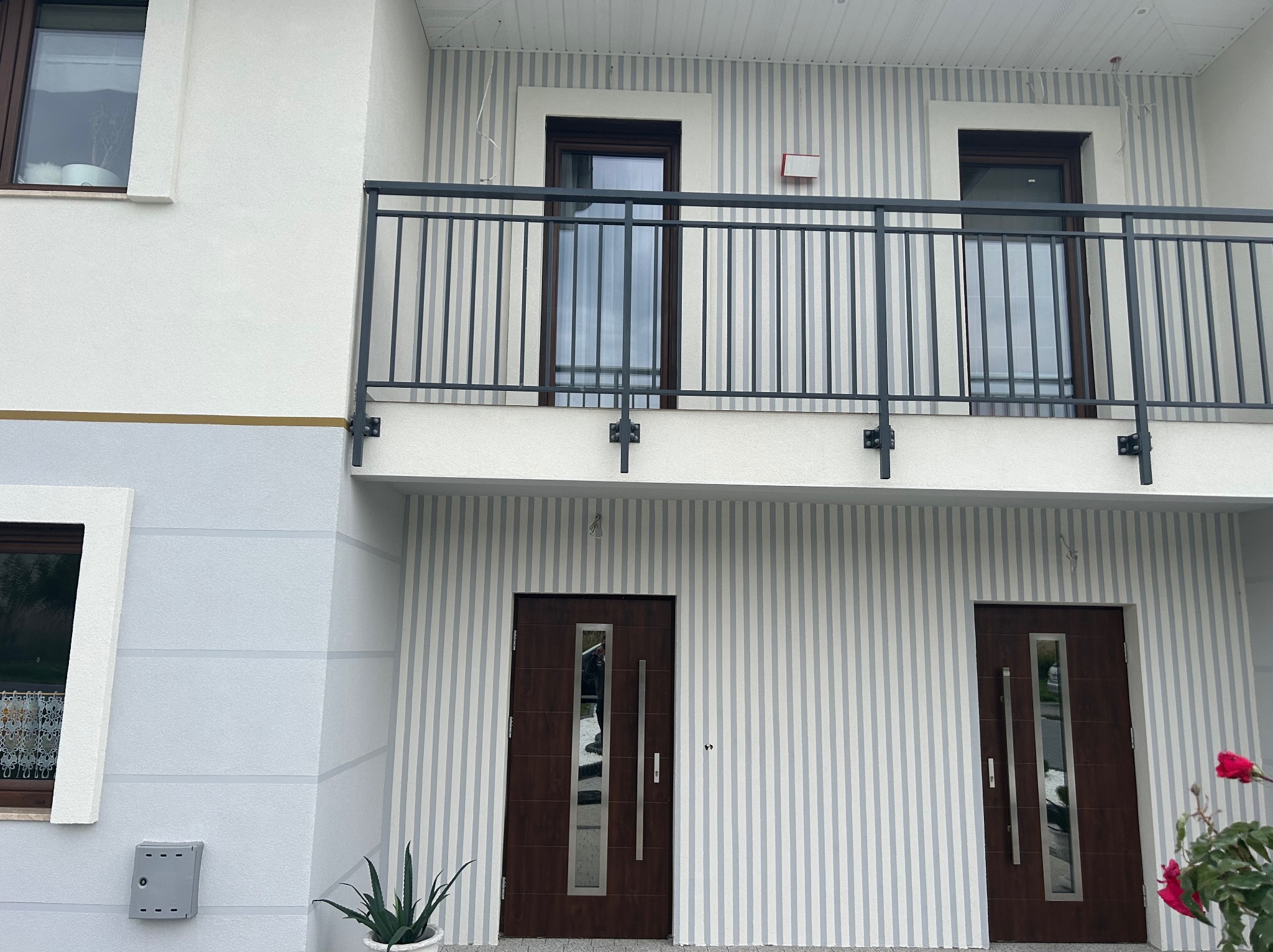 Elewacja domu z balkonem i dwoma wejściami, wykończona w pionowe pasy szarości i bieli, z ciemnymi oknami i drzwiami oraz czarną balustradą balkonu.