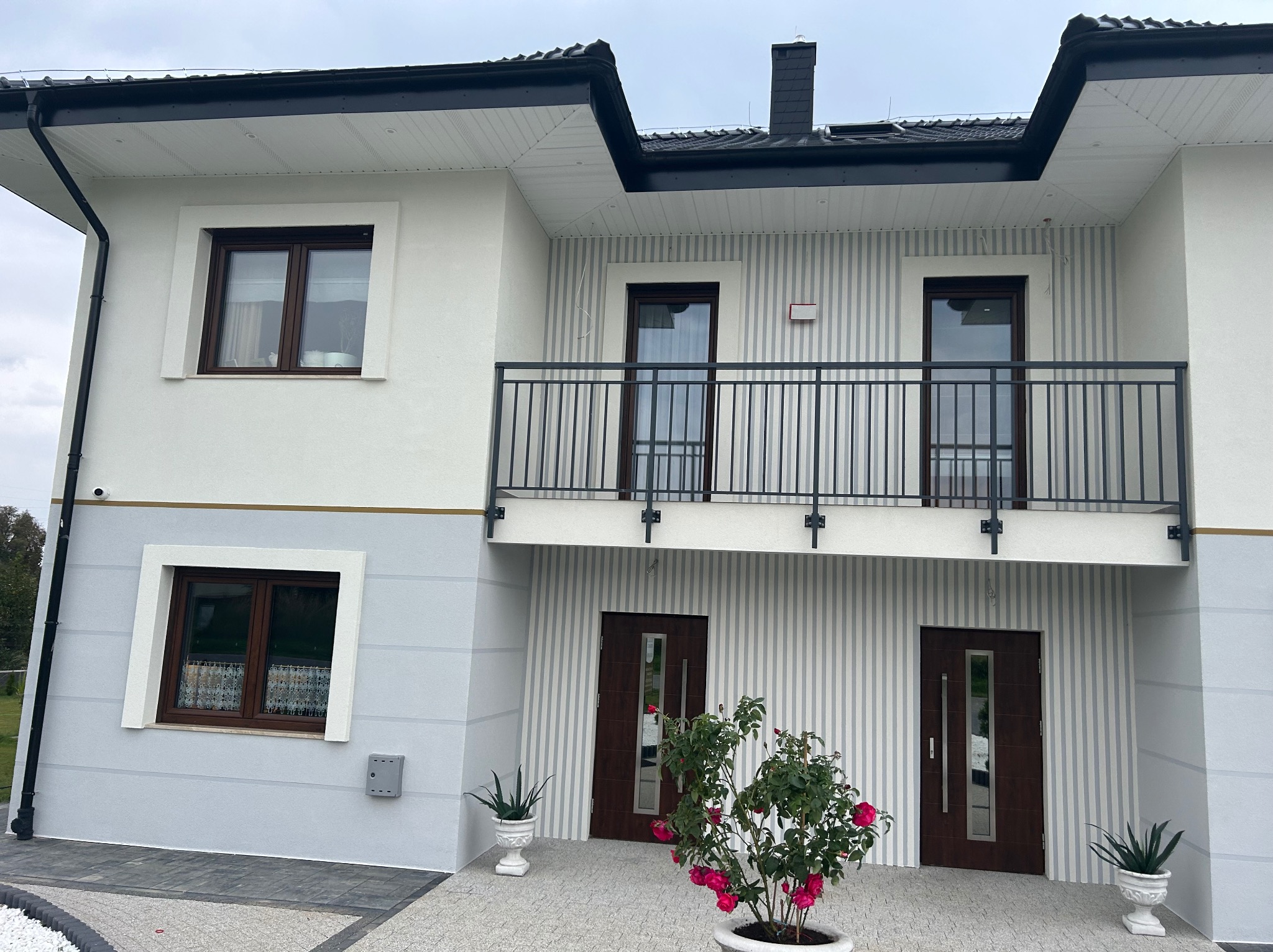 Dwupiętrowy dom z balkonem, elewacja w odcieniach szarości i bieli, z ciemnymi oknami i drzwiami, oraz kwiatami w donicach przed wejściem.