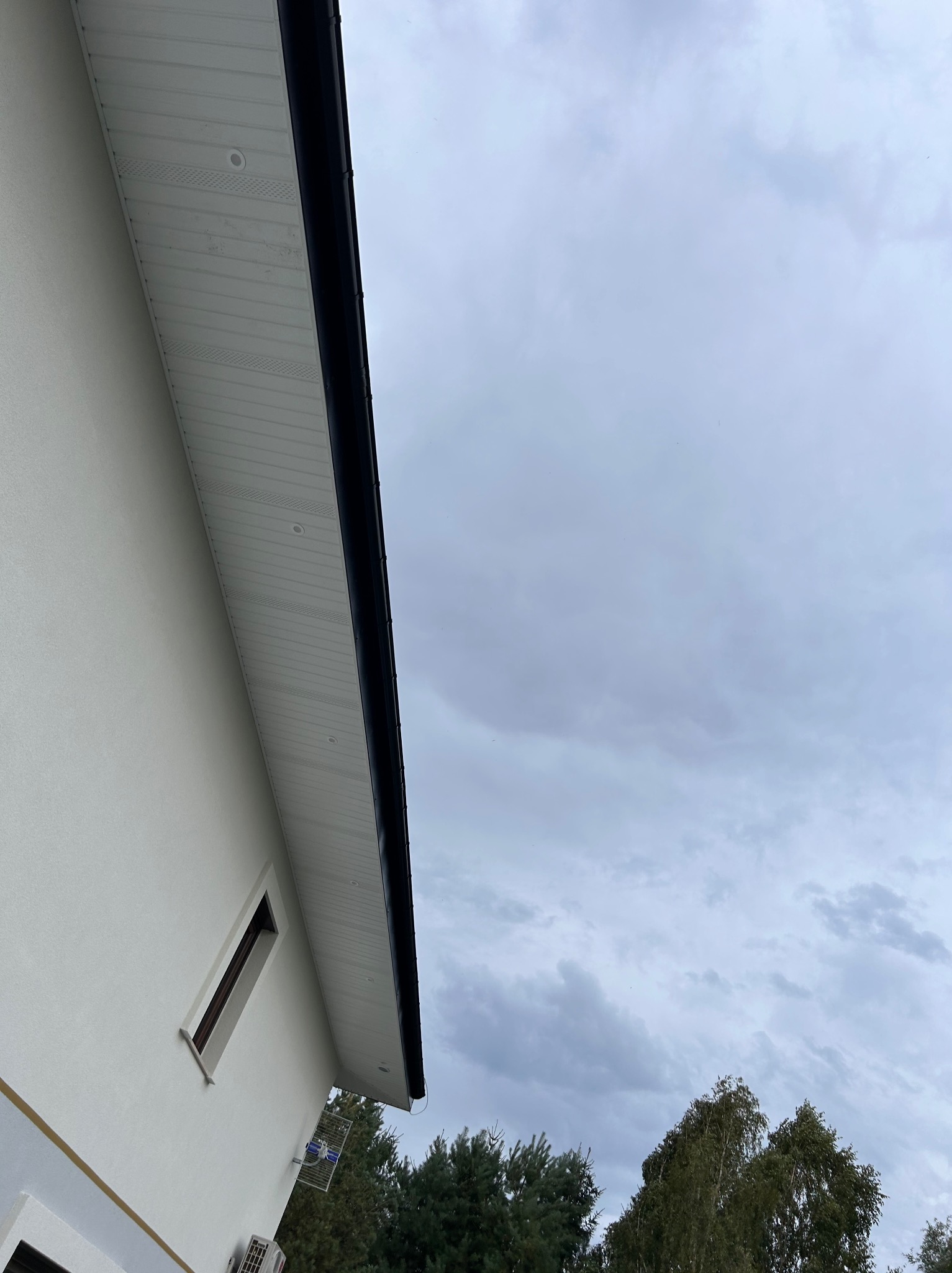 Fragment elewacji domu z białym tynkiem, ciemną rynną i podbitką z oświetleniem punktowym, widok z dołu na pochmurne niebo i drzewa.