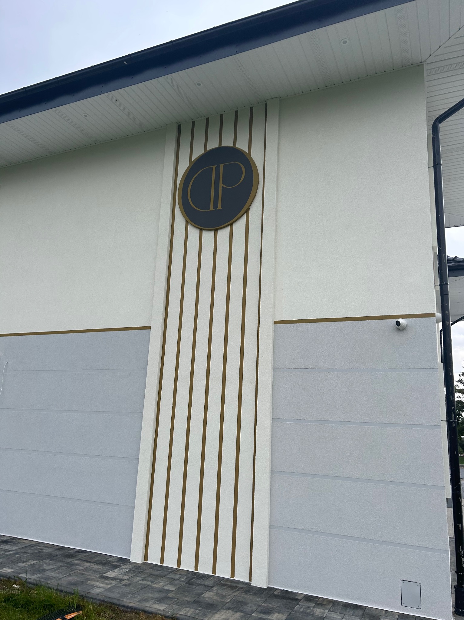 Elewacja budynku z szarym pasem i pionowymi złotymi paskami, okrągłe logo firmy i kamera monitoringu