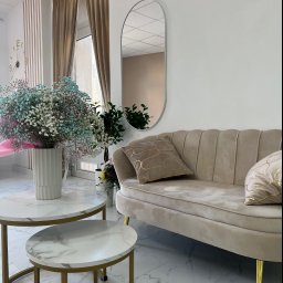 PROremont Kraków - Beżowa sofa z poduszkami w salonie, stoliki kawowe z marmurowym blatem i bukiet gipsówki w wazonie, lustro owalne na ścianie, zasłony w oknie.