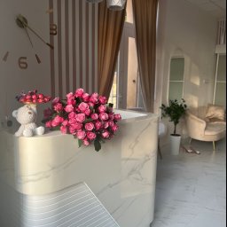 PROremont Kraków - Recepcja w salonie kosmetycznym z marmurowym blatem, dekorowana bukietem różowych róż, figurką misia trzymającego paterę z czekoladkami, zegarem ściennym i trzema nowoczesnymi lampami wiszącymi.