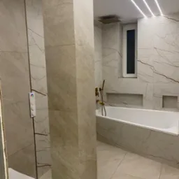 Wykończona łazienka z beżowymi płytkami imitującymi marmur, podświetlona listwami LED na suficie, z wanną, złotą armaturą i podtynkową spłuczką z kolorowym podświetleniem.