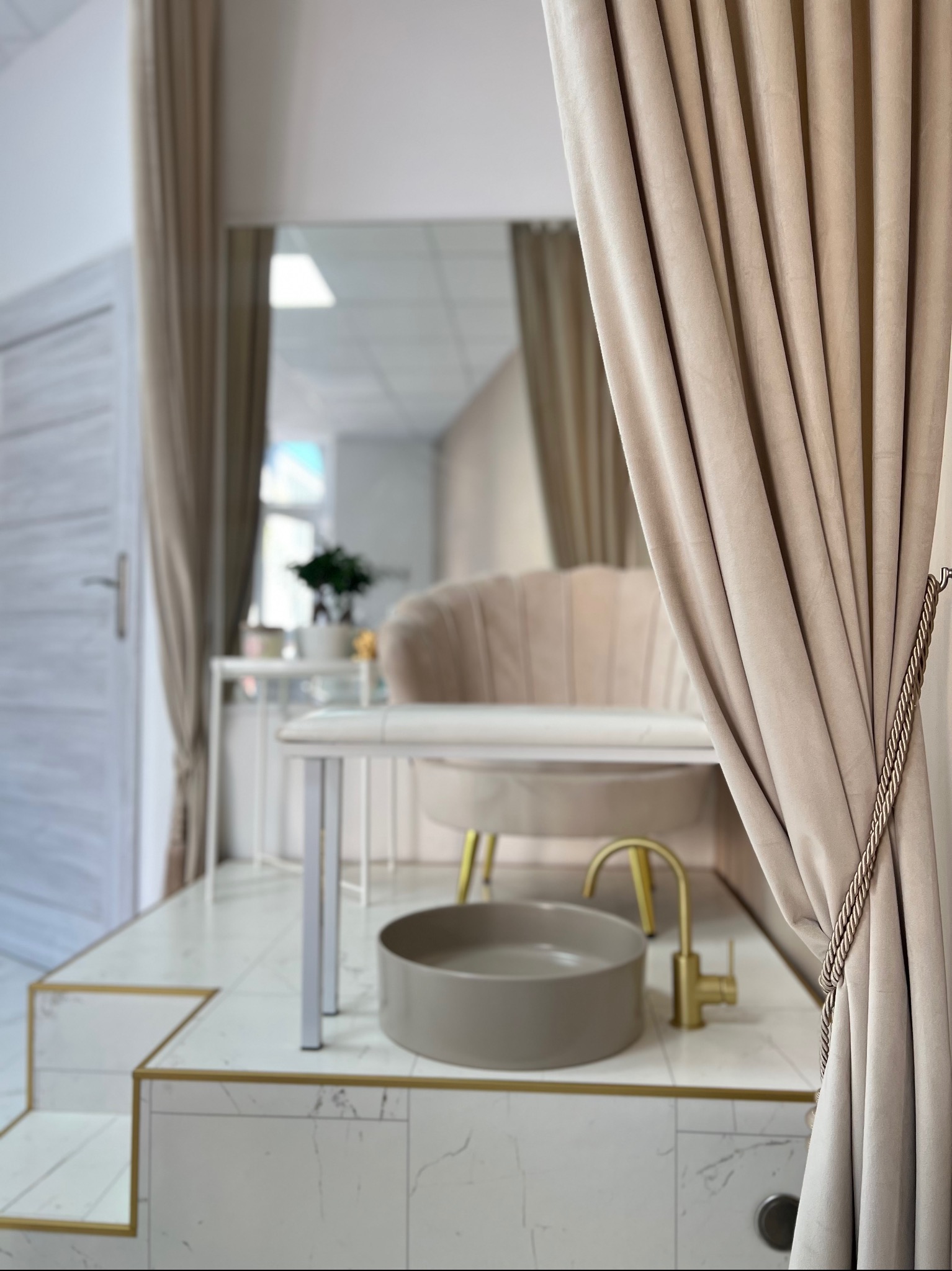 Beżowa zasłona z ozdobnym sznurem w salonie kosmetycznym; w tle marmurowe płytki, umywalka i fotel do pedicure.
