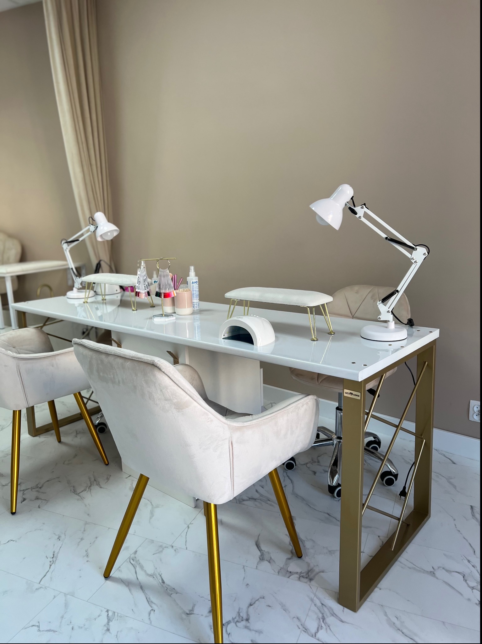 Dwa stanowiska do manicure z białymi blatami i złotymi nogami, krzesła welurowe na złotych nóżkach, lampki biurkowe, akcesoria do stylizacji paznokci, marmurowa podłoga.