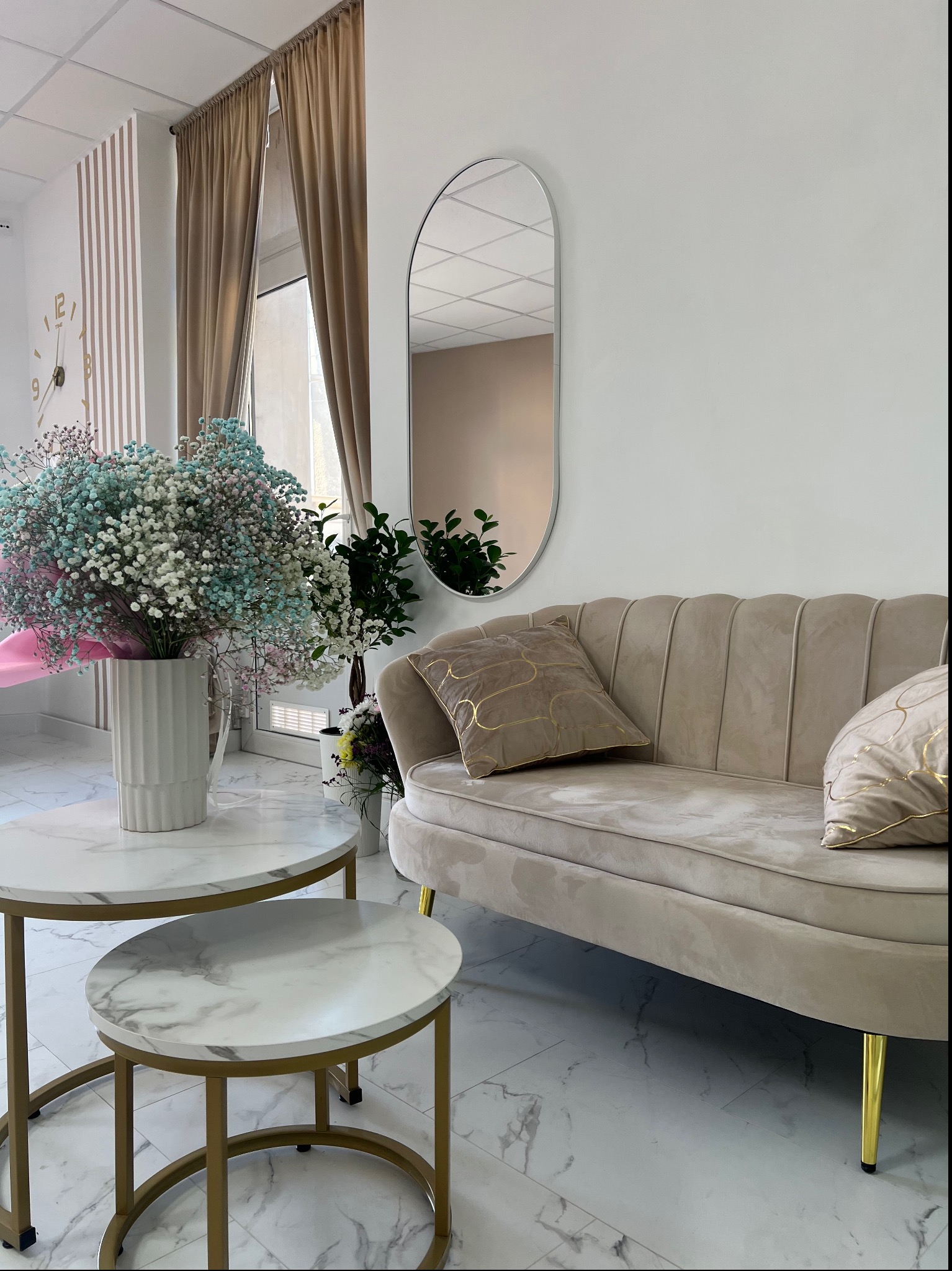 Beżowa sofa z poduszkami w salonie, stoliki kawowe z marmurowym blatem i bukiet gipsówki w wazonie, lustro owalne na ścianie, zasłony w oknie.