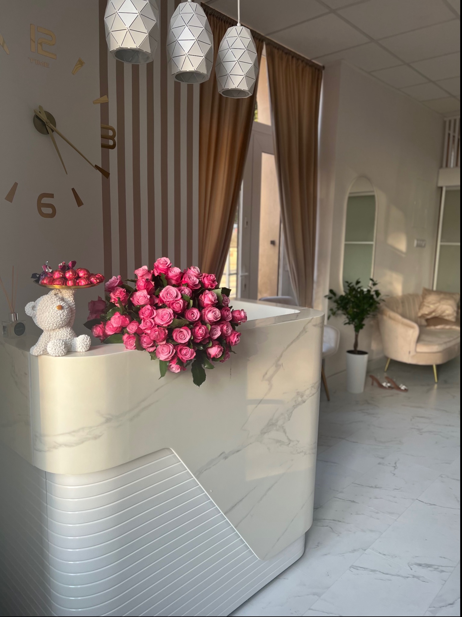 Recepcja w salonie kosmetycznym z marmurowym blatem, dekorowana bukietem różowych róż, figurką misia trzymającego paterę z czekoladkami, zegarem ściennym i trzema nowoczesnymi lampami wiszącymi.