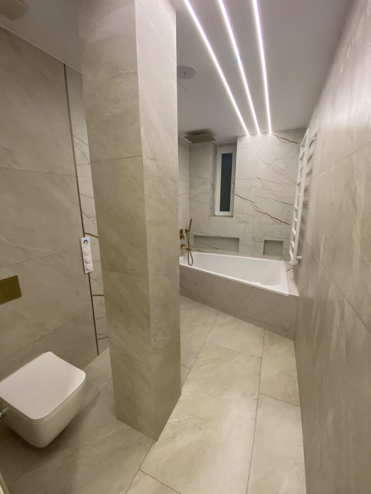 Nowoczesna łazienka wykończona w beżowych płytkach imitujących marmur, z podświetlanym sufitem, zabudowaną wanną i podwieszaną toaletą z panelem sterowania.