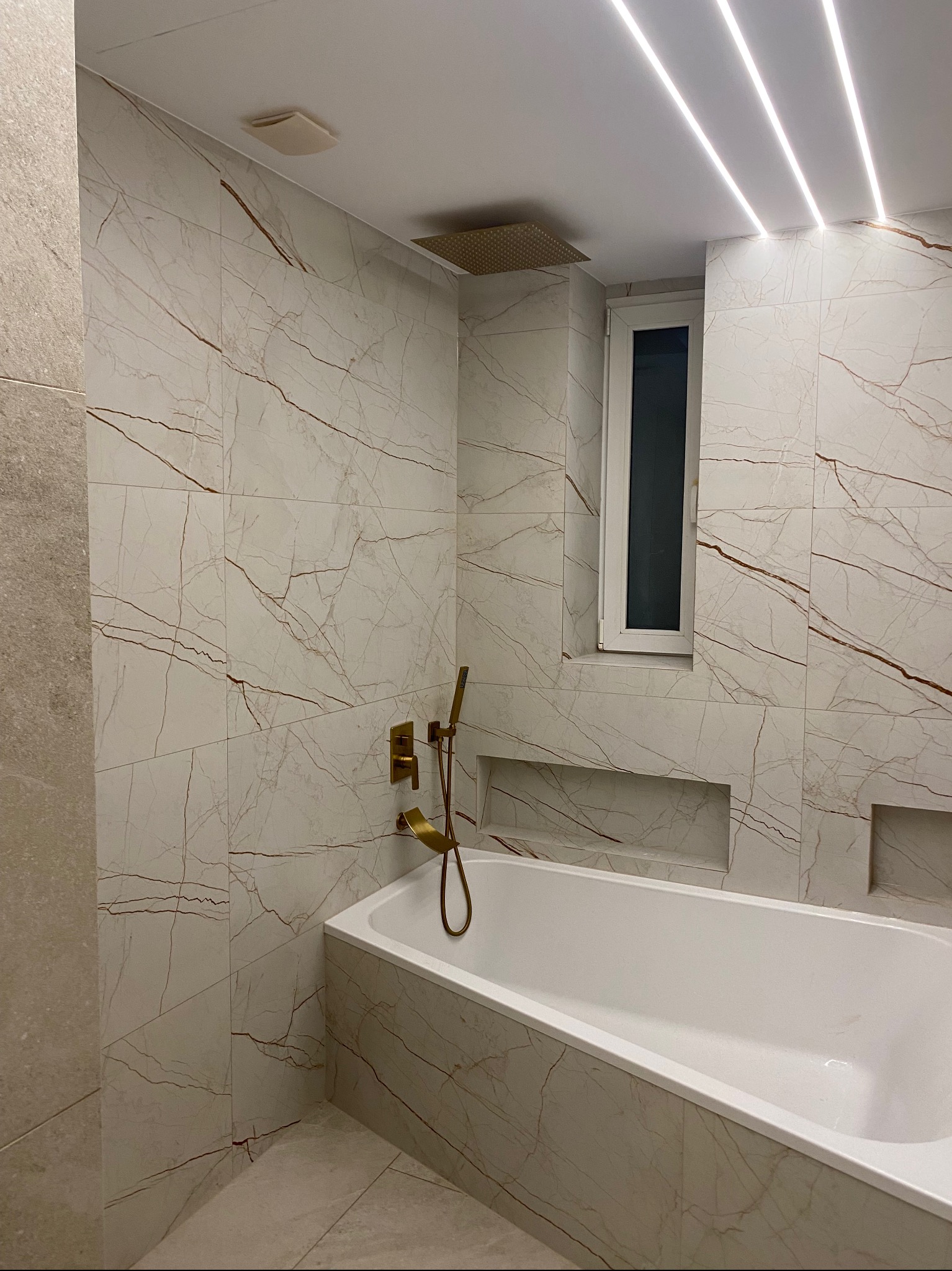 Nowoczesna łazienka z wanną obłożoną płytkami imitującymi marmur z brązowymi żyłami, złota armatura, oświetlenie LED w suficie i wnęki w ścianie.