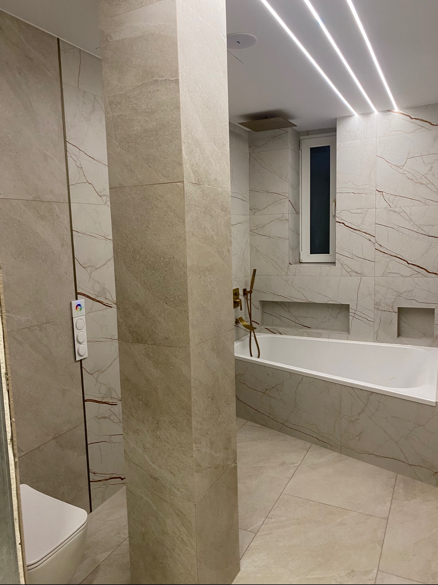 Wykończona łazienka z beżowymi płytkami imitującymi marmur, podświetlona listwami LED na suficie, z wanną, złotą armaturą i podtynkową spłuczką z kolorowym podświetleniem.