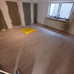 MAXI usługi wykończeniowe - Pokój w trakcie prac wykończeniowych: świeżo ułożone panele podłogowe, fragment ściany z nałożoną gładzią, żółta folia ochronna na podłodze, narzędzia na parapecie, widoczna szczotka do zamiatania.