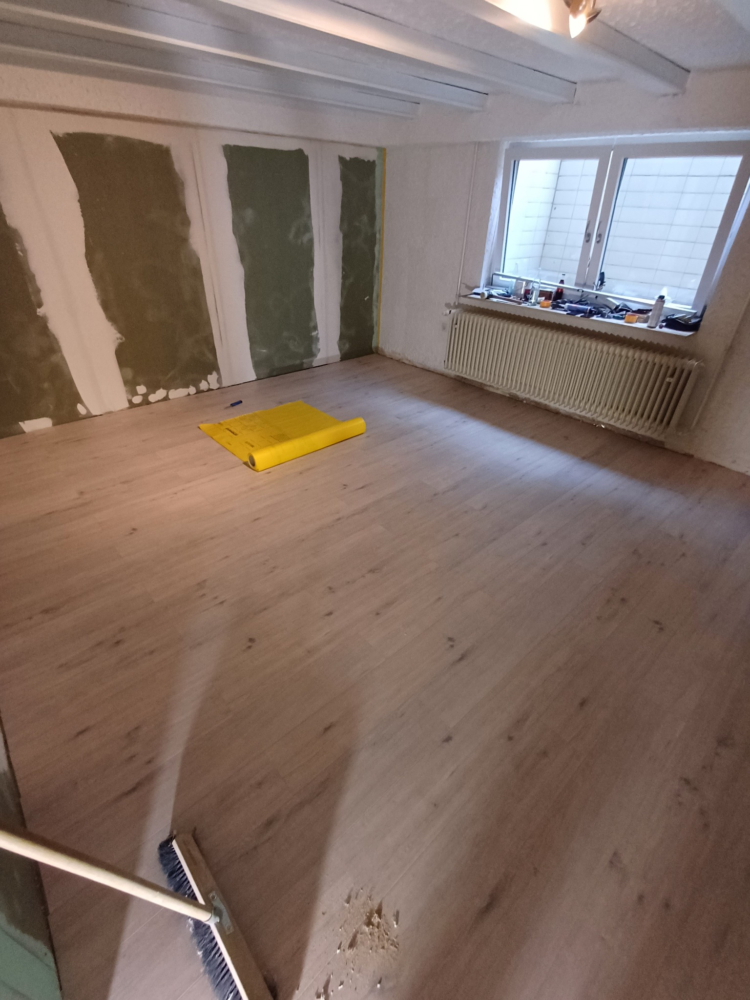 Pokój w trakcie prac wykończeniowych: świeżo ułożone panele podłogowe, fragment ściany z nałożoną gładzią, żółta folia ochronna na podłodze, narzędzia na parapecie, widoczna szczotka do zamiatania.