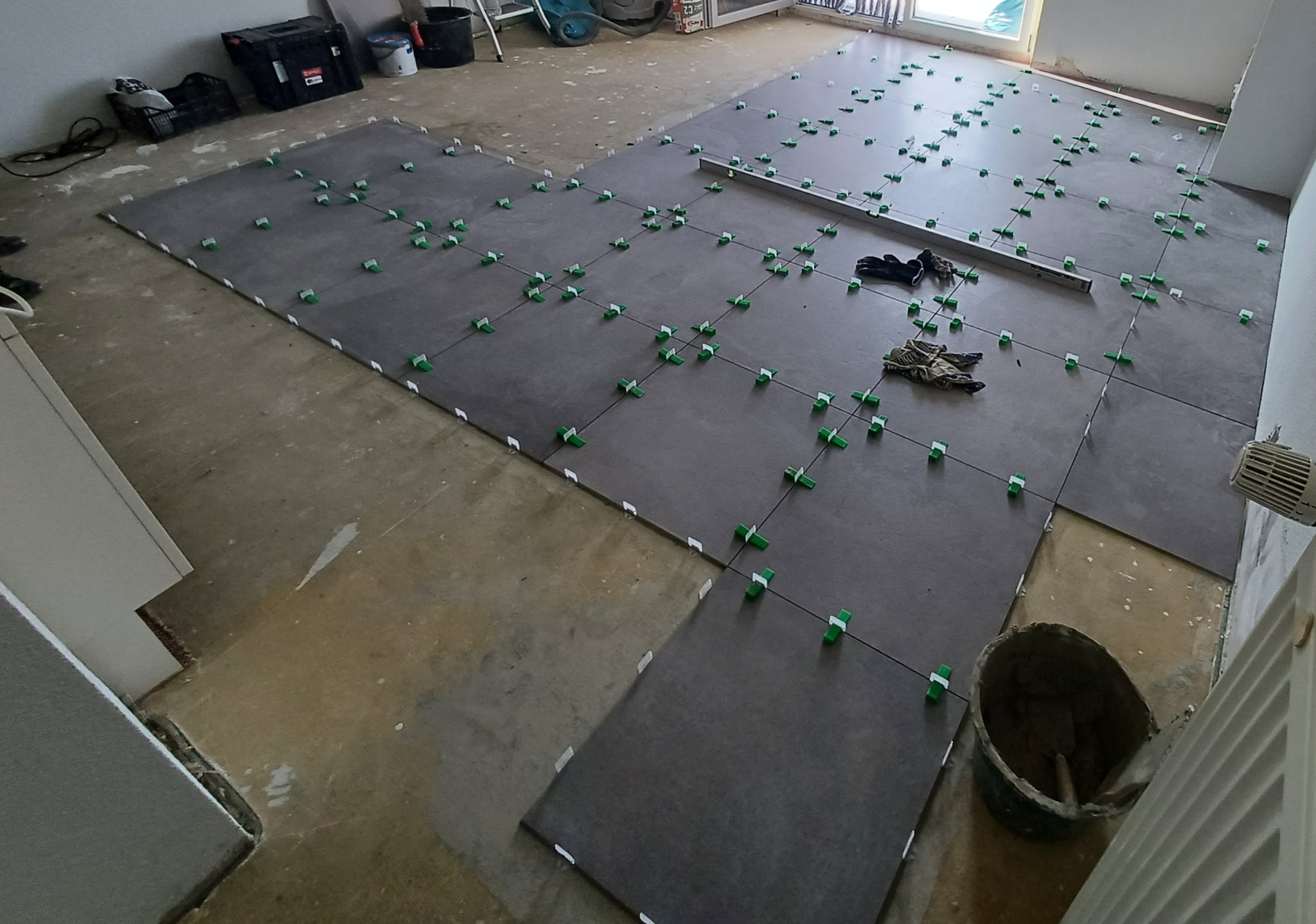 Układanie szarych płytek podłogowych z użyciem zielonych klipsów poziomujących, widoczna poziomica, wiadro z zaprawą i rękawice robocze.