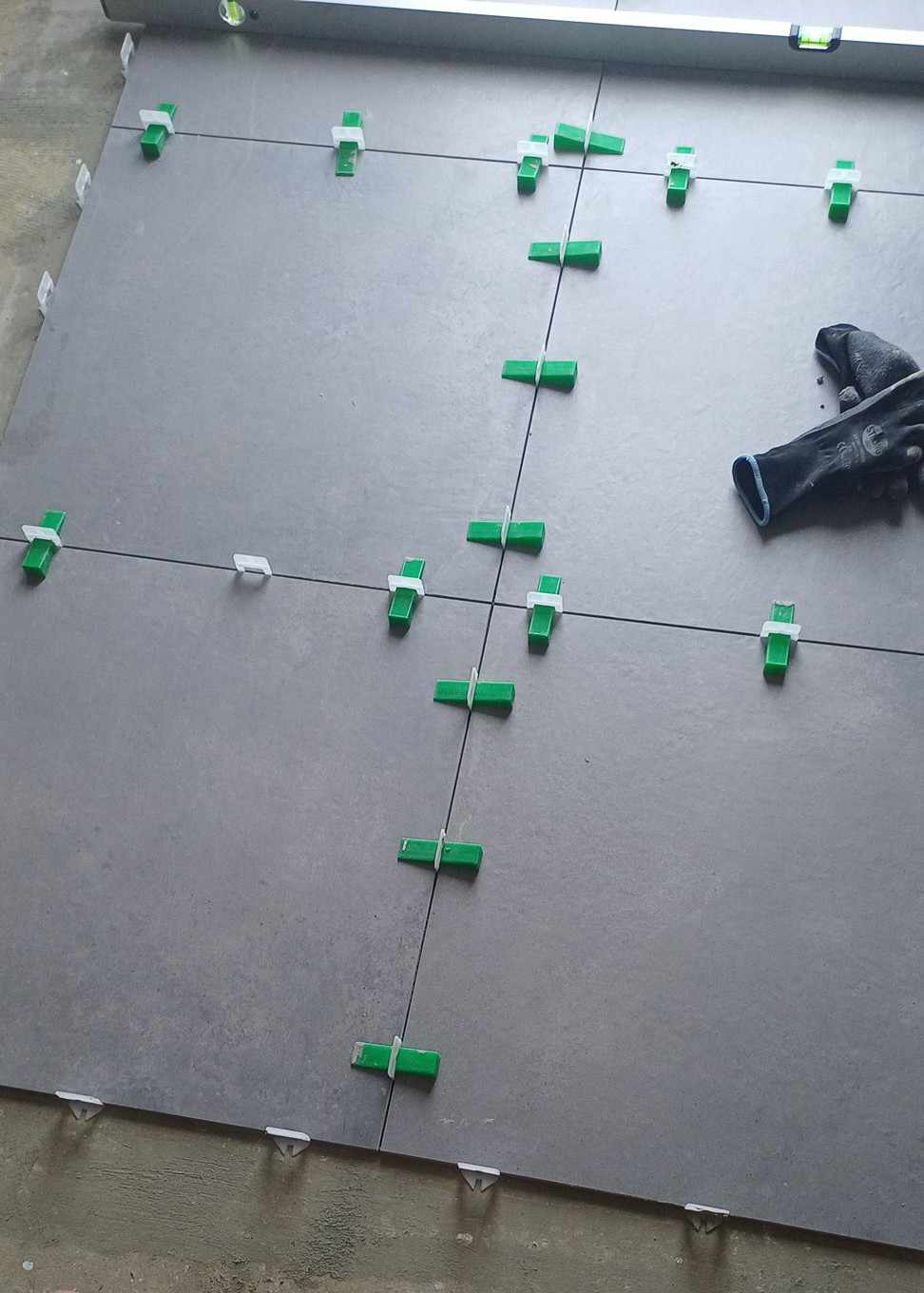 Układanie szarych płytek z użyciem poziomicy i zielonych klipsów poziomujących, widoczna czarna rękawica robocza.