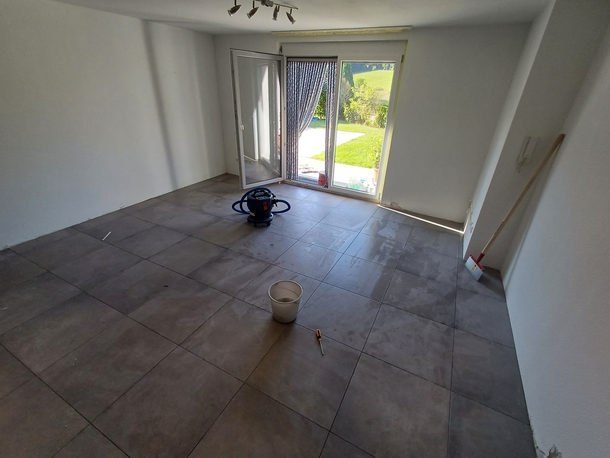 Pokój po remoncie z szarymi płytkami podłogowymi, otwartym oknem balkonowym z widokiem na ogród, odkurzaczem, wiadrem, śrubokrętem i szpachelką opartą o ścianę.