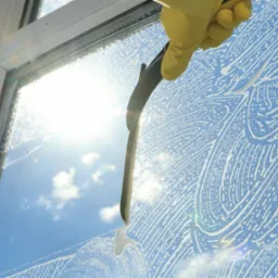 Okno z widocznym niebem i chmurami podczas czyszczenia za pomocą ściągaczki i mydlin, ręka w żółtej rękawicy w górnym rogu.