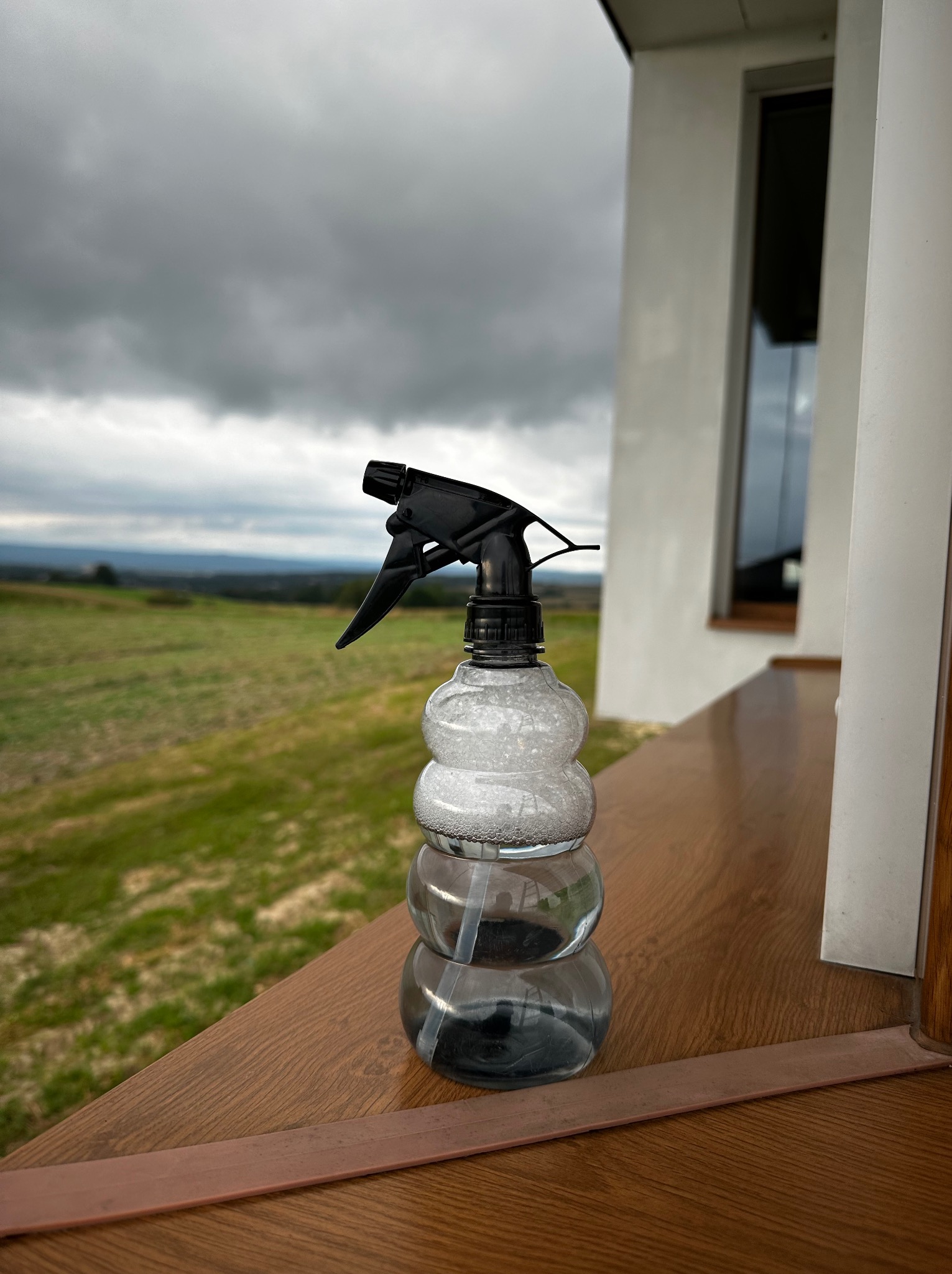 Plastikowy spryskiwacz z płynem na drewnianym parapecie z widokiem na zielone wzgórza i pochmurne niebo w tle.