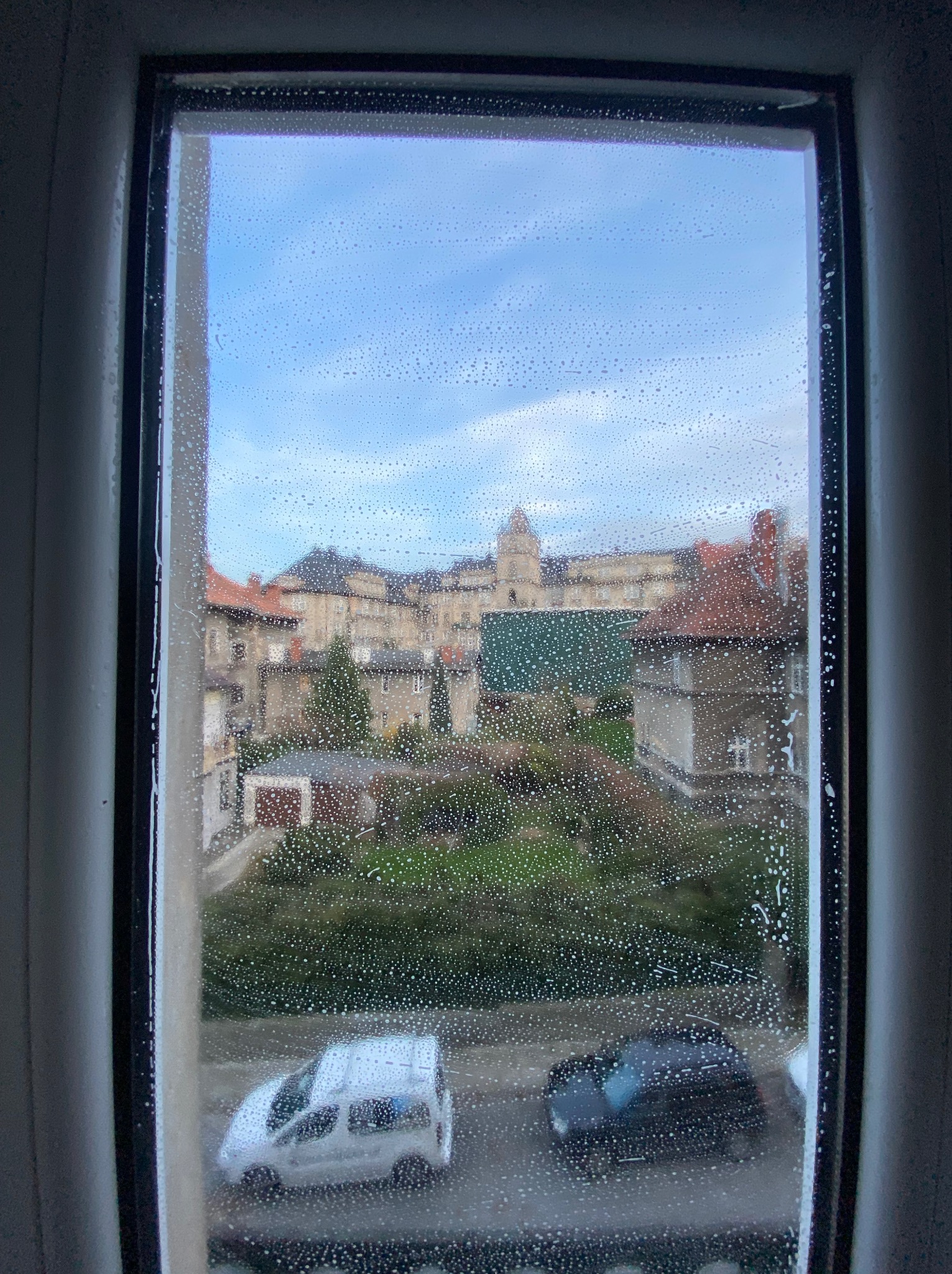 Okno w trakcie mycia, z widocznymi kroplami wody i piany, za oknem widok na budynki mieszkalne i dwa zaparkowane samochody.