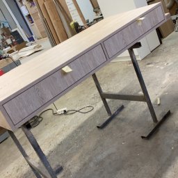 AG-MAR - Biurko z szufladami w trakcie produkcji, z blatem imitującym drewno, ustawione na metalowych nogach w warsztacie stolarskim.