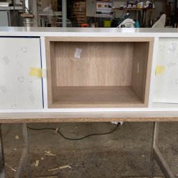 AG-MAR - Niedokończona szafka z białymi frontami zabezpieczonymi folią ochronną i otwartą wnęką z jasnego drewna, umieszczona na metalowym stojaku w warsztacie.