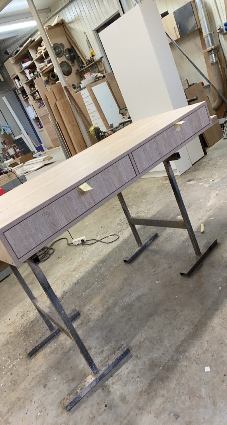 Biurko z szufladami w trakcie produkcji, z blatem imitującym drewno, ustawione na metalowych nogach w warsztacie stolarskim.