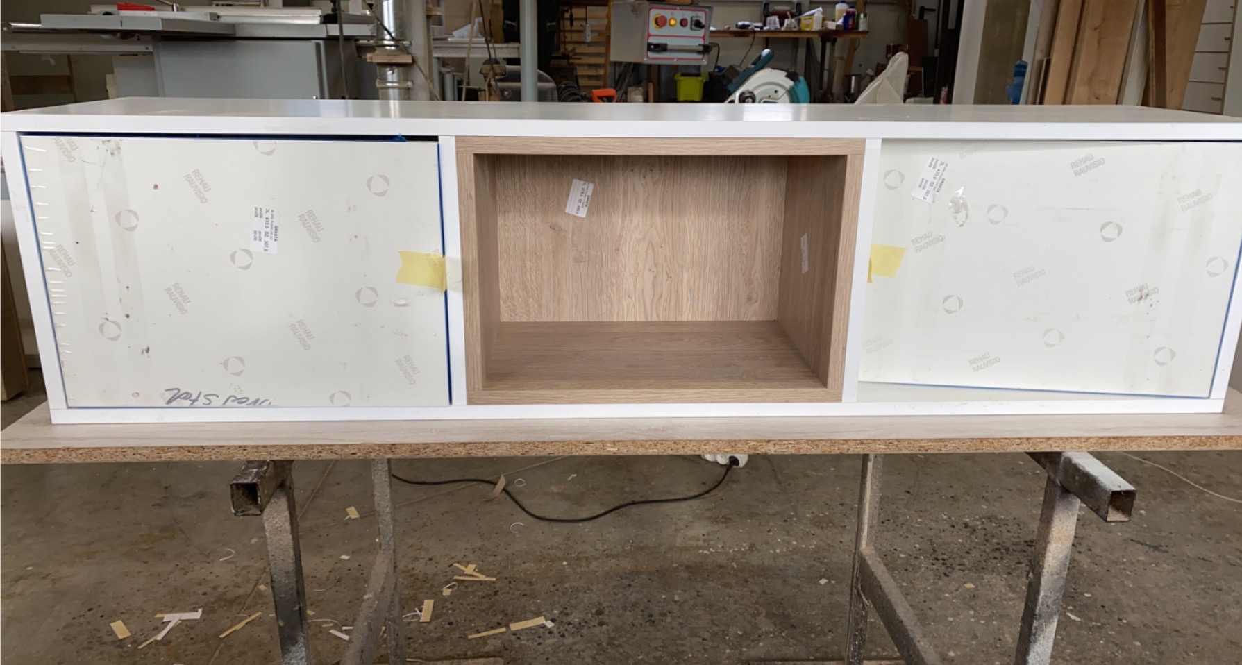 Niedokończona szafka z białymi frontami zabezpieczonymi folią ochronną i otwartą wnęką z jasnego drewna, umieszczona na metalowym stojaku w warsztacie.