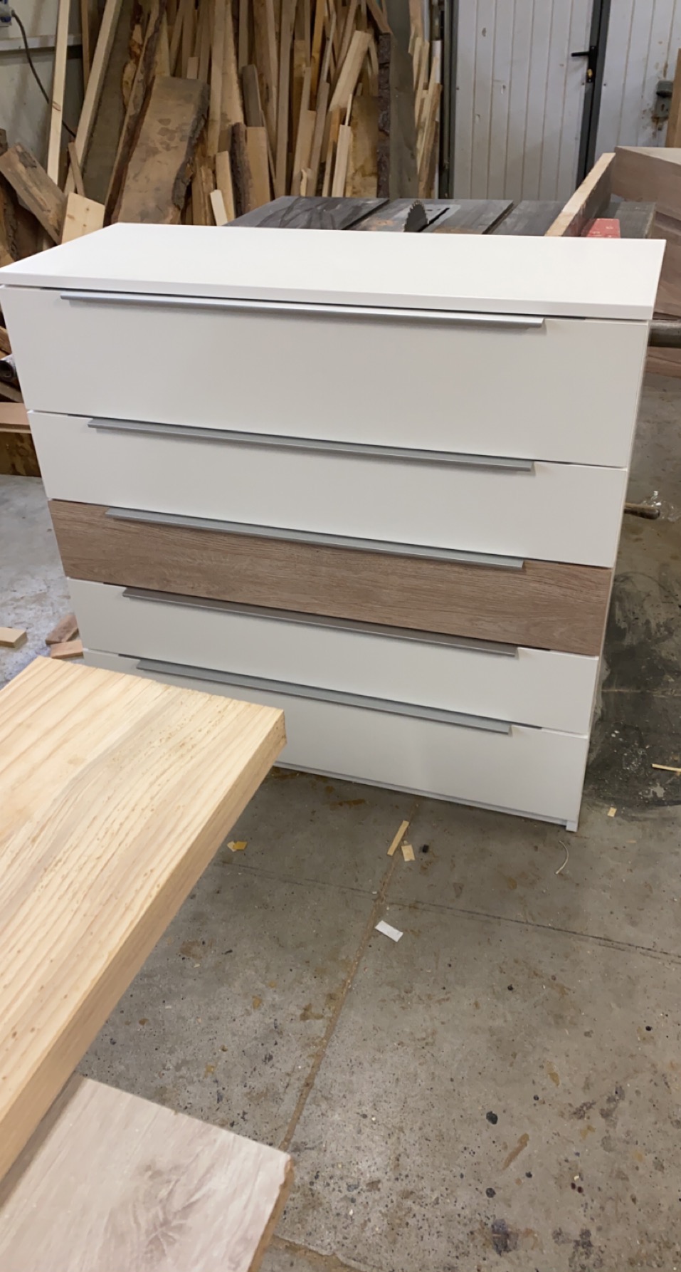 Biała komoda z czterema szufladami, z jedną szufladą w kolorze drewna, w warsztacie w Morawicy, z widocznymi deskami w tle.